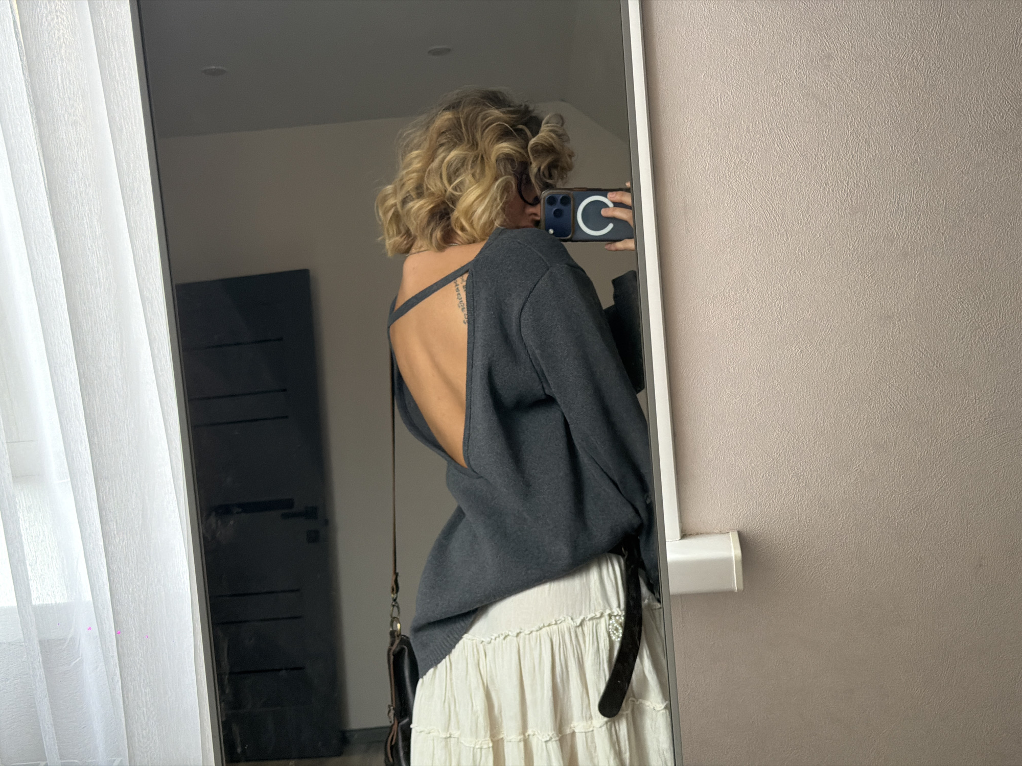 A woman with curly blonde hair stands in front of a mirror, taking a selfie with her phone. She is wearing a dark grey, open-back sweater over a cream-colored tiered skirt. A dark leather purse with a strap is slung over her shoulder. The background of the mirror reflection shows a white curtain on the left and a dark door on the right. The wall in front of the mirror is a textured, light brown. The location is in the city of Kamenka, Russia.