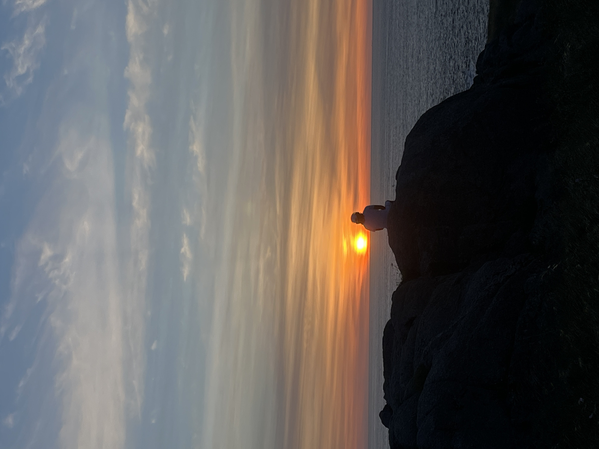 A horizontal outdoor shot depicts a sunset at an unknown location on the coast of Spain. The sky transitions from light blue with white streaks in the upper-left, through orange, to darker shades of orange and red near the horizon, where the setting sun shines brightly. A person is silhouetted against the sun on top of a rocky outcrop, which is dark and jagged. The sea stretches horizontally along the right edge of the frame, with a calm surface reflecting the colors of the sunset. The image is oriented sideways, with the sky and sea appearing to run vertically.
