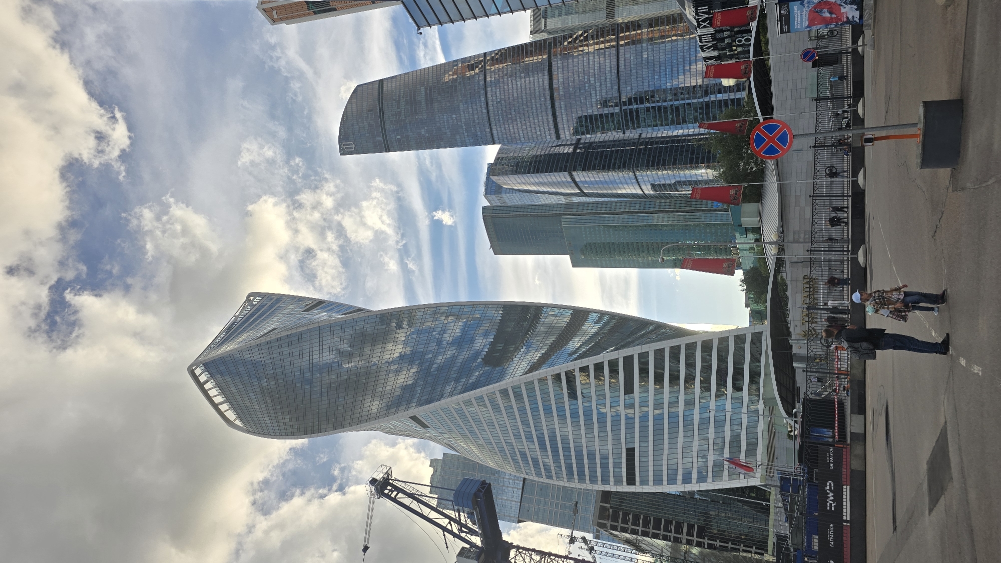 The image shows a wide-angle view of the Moscow International Business Center (also known as Moscow-City) in Moscow, Russia. The scene is dominated by several modern skyscrapers, including the distinctive twisted skyscraper "Neva Towers" and the rounded skyscrapers "Eurasia" and "Capital City." The sky is partly cloudy with dramatic clouds reflecting on the glass facades of the buildings.
In the foreground, on an asphalt road, a man wearing a dark jacket, blue jeans, and a backpack is standing. Beside him, a child wearing a plaid shirt, shorts, and a light blue cap is walking. They are facing away from the camera, looking towards the skyscrapers.
Behind them, a black metal fence encloses a pedestrian area with several people walking. Red flags with white lettering are displayed along the fence. A traffic sign with a blue circle and red cross is visible on the right side of the road, indicating no stopping or parking. Construction cranes are visible to the left of the frame, suggesting ongoing development in the area. The overall impression is that of a bustling urban environment with a mix of modern architecture and everyday life.