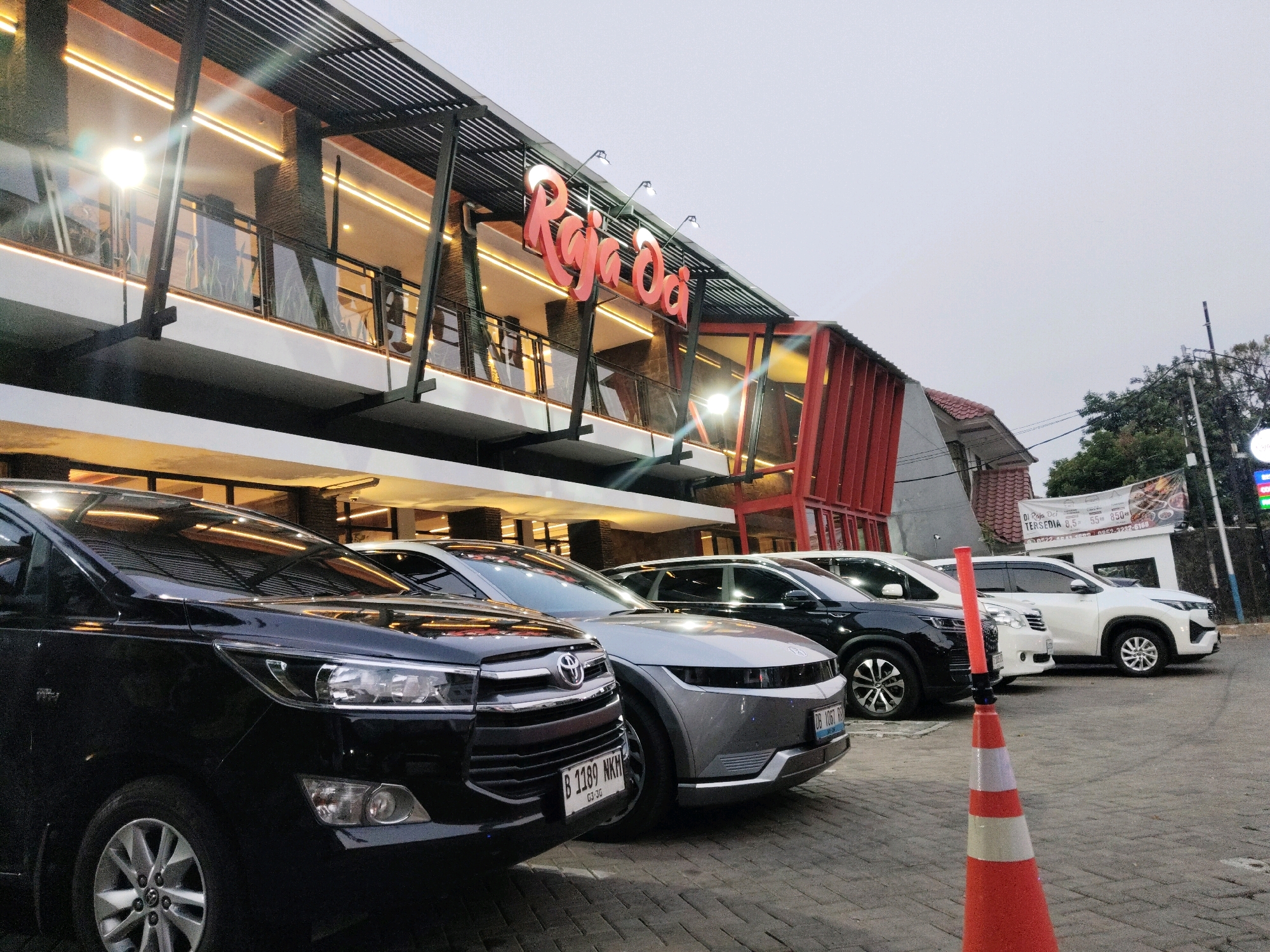 The image shows a row of cars parked in front of a building with a sign that says "Raja Ja Da". The building appears to be a restaurant or a commercial establishment, with modern architecture featuring glass windows, a dark metal roof structure, and red accents. The cars are parked on a paved surface, possibly a parking lot, with a cobblestone-like pattern. The sky is overcast, suggesting it might be evening or a cloudy day. A traffic cone is visible in the foreground, suggesting ongoing construction or a temporary traffic management situation. The cars appear to be a black SUV, a silver electric vehicle, a black SUV, and a white SUV, lined up in a parking area. There are no people visible in the image.