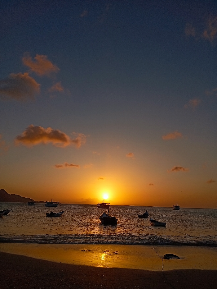Sunset view of the ocean. The sun is positioned above the horizon, with several boats dotting the water. The clouds are lightly colored in hues of orange and pink. The beach appears in the foreground with reflections mirroring the sun.