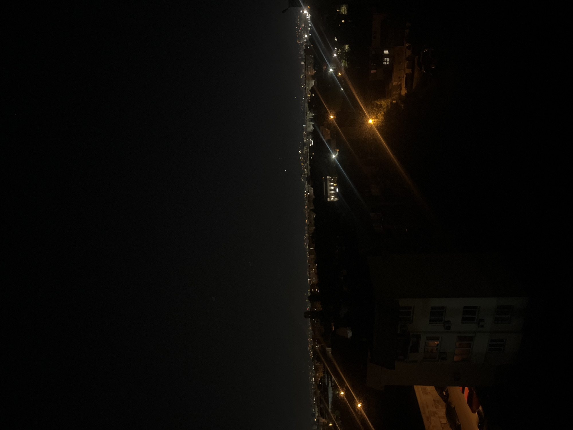 The image depicts a nighttime scene in what appears to be a coastal city, possibly near Sofia, Bulgaria. It is oriented vertically, with the coastline running along the center from top to bottom.
On the left side is a dark expanse, likely the sea or a large body of water. On the right, there is a city illuminated by streetlights and building lights. The lights extend along the coastline, indicating a developed urban area. Some buildings are partially visible, including multi-story structures with lighted windows. Streetlights are positioned along the road, creating streaks of light extending into the distance. At the bottom of the image are objects near the viewer, like an overhanging building and a street.