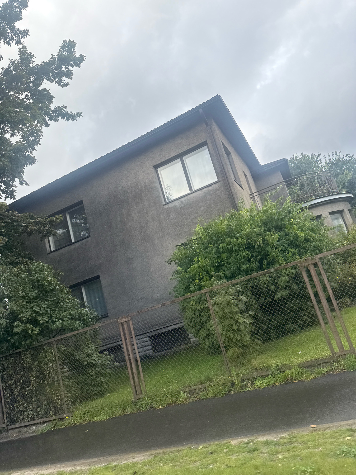 The image shows a two-story grey concrete house with a dark roof. The house has several windows with dark frames. There is a curved balcony on the right side of the house, adorned with an ornate metal railing. Lush green bushes and trees surround the house, with some branches from a large tree extending from the left side of the frame. A chain-link fence with metal posts runs across the foreground, separating the property from a paved walkway and a grassy area. The sky is overcast with grey clouds. This scene is located in the Pirita district of Tallinn, Estonia. There are no people visible in the image.