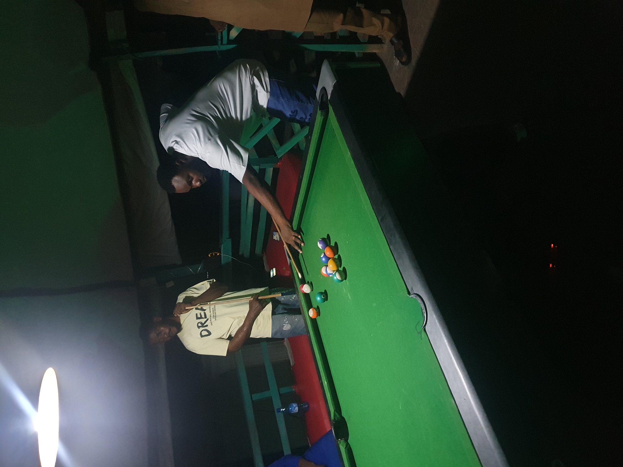 In Lokoja, Nigeria, two men are playing pool. The man on the right, wearing a light-colored t-shirt and blue shorts, is in the process of striking a billiard ball with a cue stick. His body is angled towards the table, and his gaze is focused on the game. To his left, another man, wearing a light t-shirt with the word "DREAM" printed on it, stands with a cue stick in his hand, observing the game. A green pool table with several colorful balls is situated between them, illuminated by an overhead light fixture. The surrounding environment appears to be an outdoor setting, possibly a bar or recreational area, with a red-colored surface visible beneath the pool table and green railings in the background. The lighting suggests it is nighttime.