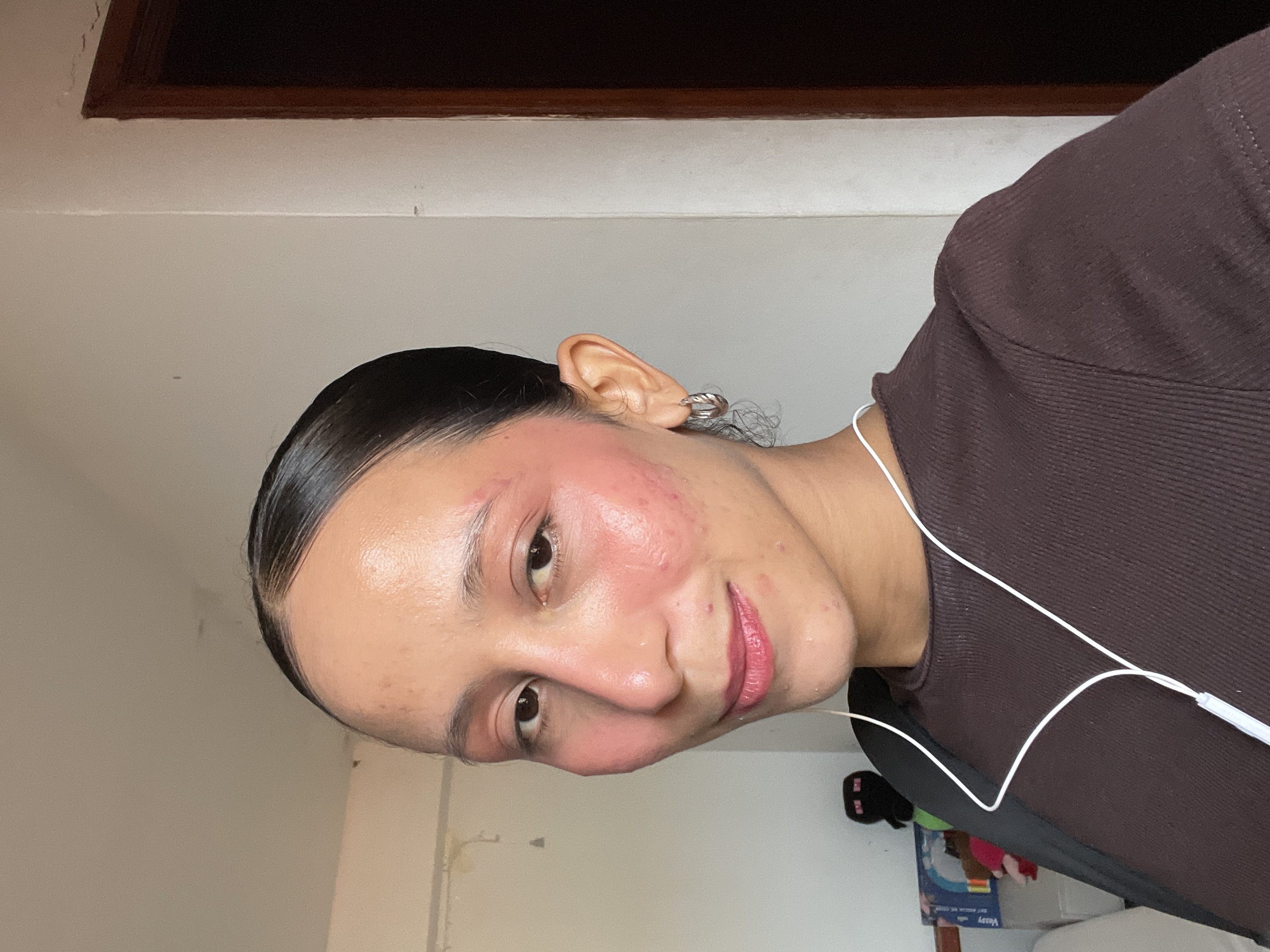 A person is shown from the chest up, looking directly at the camera with a slight smile. They have dark hair pulled back tightly, revealing a high forehead. Their skin has a warm tone and appears to have a dewy finish. There is noticeable pink blush applied to their cheeks and extending up to the temples and over the eyelids. Small blemishes are visible on the face. The person is wearing a dark brown, ribbed top. White earbuds with a cord are visible, with the cord running down the front of their shirt. They are wearing a small silver hoop earring in their visible ear. The background consists of a plain, light-colored wall. To the left of the person, a shelf with various items, including stuffed toys and what appears to be a Minecraft character, is partially visible. To the right, a portion of a dark brown wooden doorframe is present. The lighting suggests an indoor setting, possibly a home environment in the city of Porlamar, Venezuela, given the context provided.