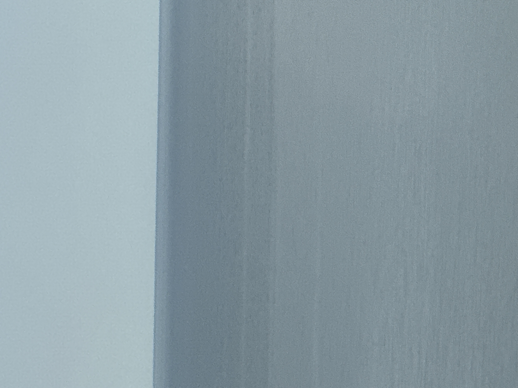 The image shows a calm, overcast sea stretching to the horizon under a pale blue sky. The water is a muted gray-blue, with subtle horizontal lines indicating ripples or currents. The sky is a uniform, soft blue. The image appears to have been taken from a high vantage point, possibly a cliff or elevated structure, overlooking the sea. The location is identified as Kholmsk, Russia. There are no people, animals, or specific structures visible in this expansive, minimalist seascape.