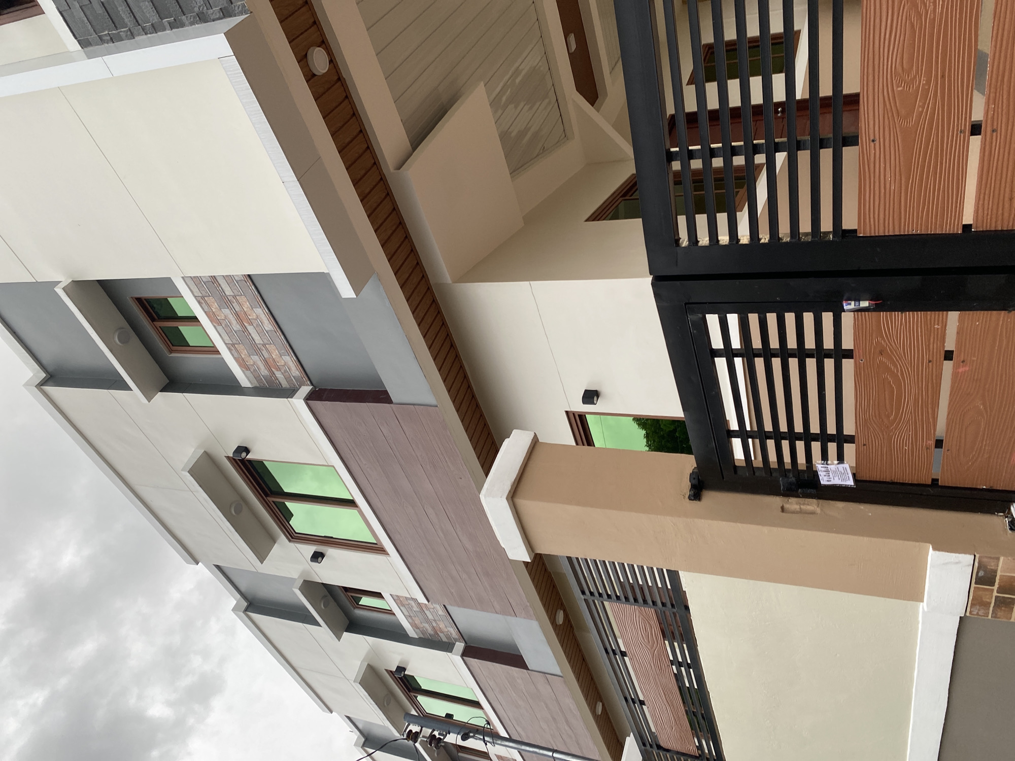 This image captures a modern multi-story residential building situated in Davao City, Philippines, under an overcast sky. The building facade is primarily painted in a light cream or off-white color, contrasted with dark grey accent sections and pronounced white architectural trim and ledges.
The structure features at least three visible levels, with multiple rectangular windows fitted with distinctive green-tinted glass and dark brown frames. Exterior wall-mounted cylindrical lights, black in color, are affixed beside several windows. Sections of the facade, particularly on the middle and lower levels, are adorned with horizontal panels featuring a reddish-brown wood grain texture, likely cement board or a similar material. Another section on the right side of the building incorporates layered stone cladding in various shades of brown, grey, and white. Overhangs and soffits beneath the upper floors and at the entrance are finished with light brown wood paneling, featuring recessed circular ceiling lights.
At ground level, a black metal gate with horizontal slats is closed, providing access to the property. The lower portions of the gate incorporate the same reddish-brown wood-grain textured panels seen on the building. A small white paper notice or sticker is affixed to the right gate panel, and a blue LED light, possibly for an access control system, is visible on the left panel. The gate is flanked on the right by a substantial tan-colored pillar with a white cap and a base clad in brick-like tiles. To the left of the pillar, a perimeter wall matching the light cream of the building is topped with a black metal fence, also featuring the reddish-brown wood-grain panels. A grey utility pole with various electrical wires and insulators is partially visible on the far left. No people or vehicles are present in the scene. The overall lighting is diffused due to the heavy cloud cover.