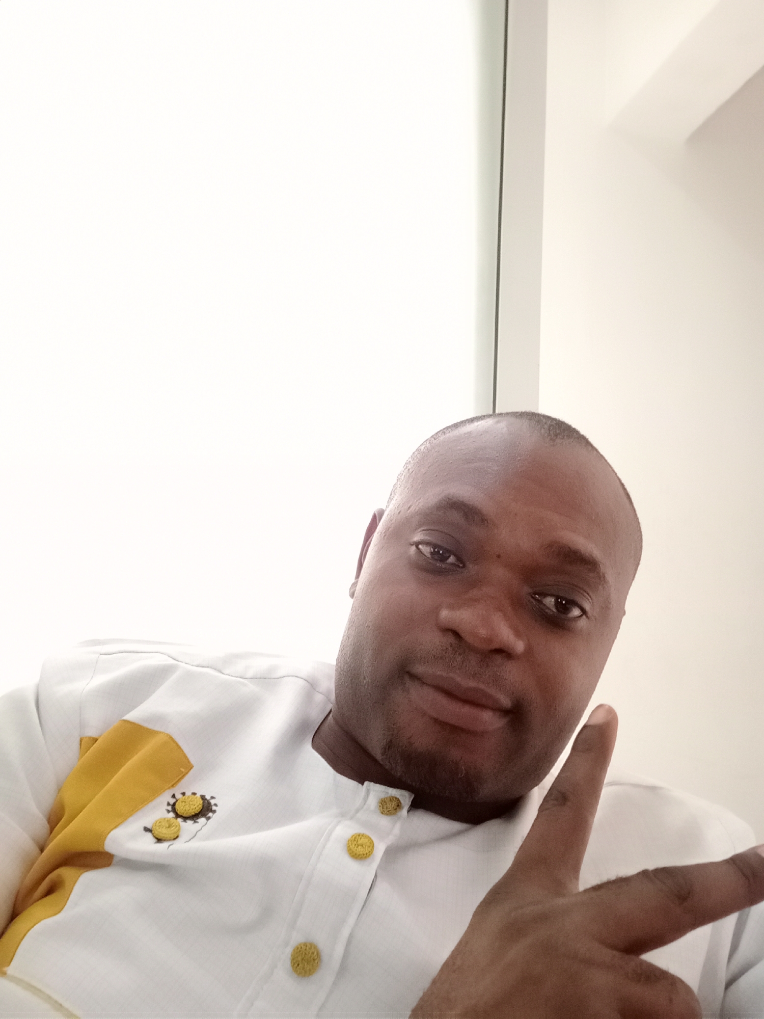 An adult man is featured in a close-up, eye-level indoor selfie or photograph, looking directly at the camera. He has dark skin, a shaved head, and a short beard or stubble. He is wearing a white long-sleeved shirt with a structured collar. The shirt features distinctive gold/yellow fabric accents on the left shoulder and chest area, along with small, circular, woven gold/yellow buttons on the placket and collar. Near the yellow fabric on the chest, there are subtle embroidered details that resemble abstract suns or gears in black and yellow.
The man's right hand is raised, with his index and middle fingers extended in a "peace" or "victory" sign, while his other fingers are curled. His expression is neutral, with a hint of a gentle smile.
The background is predominantly bright white, indicating an indoor setting with clean, minimalist walls. A thin, dark vertical line is visible on the left side, possibly part of a doorframe or window edge. The lighting is even and bright, suggesting good indoor illumination. No text is visible in the image. The location context provided is Gwarinpa, Nigeria.