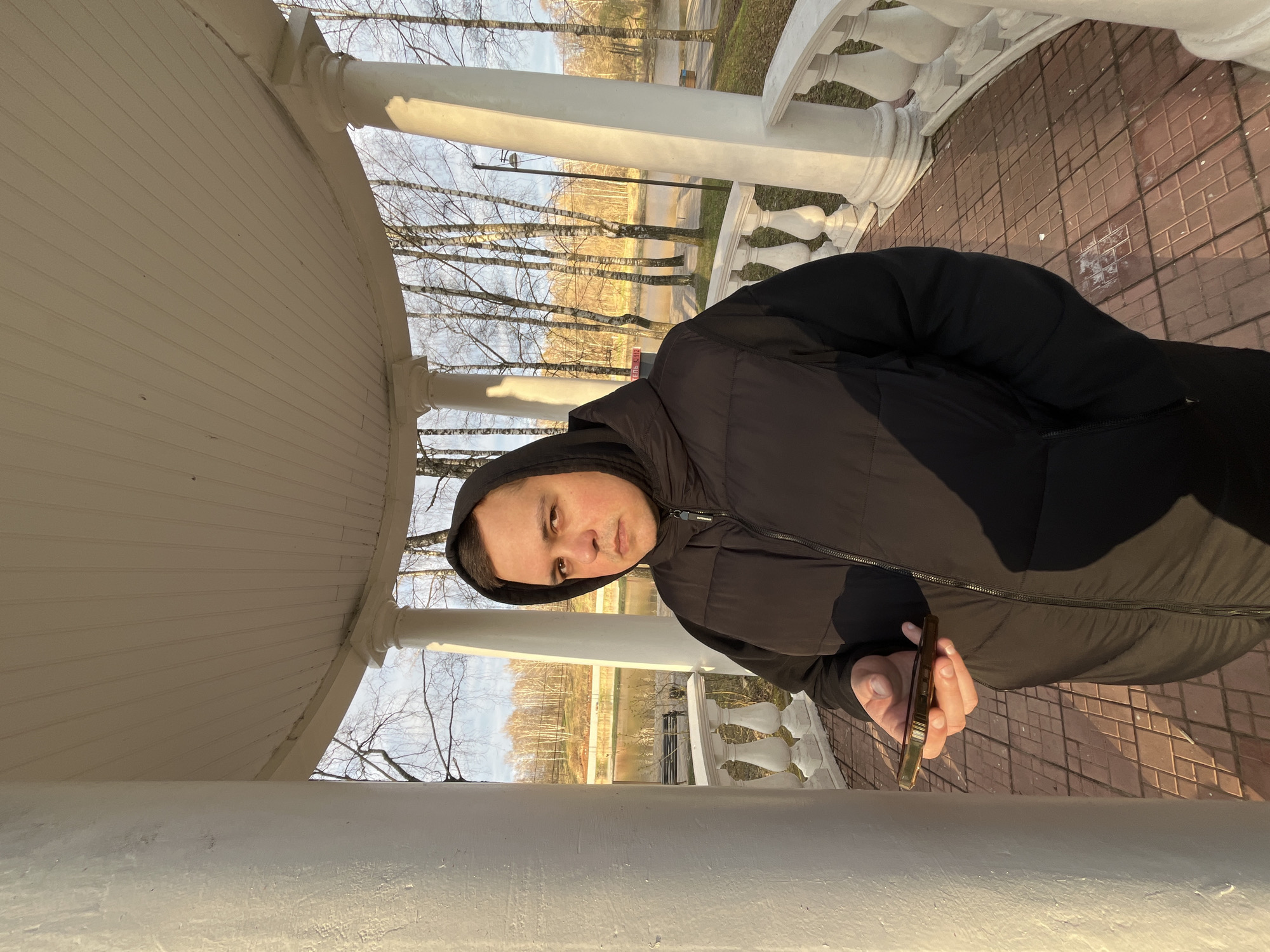 A man wearing a black hooded jacket stands inside a white gazebo-like structure with a curved roof and decorative balustrade. He holds a gold-colored mobile phone in his right hand. The ground within the structure is paved with brown tiles. Through the balustrade, trees and a pond are visible, suggesting an outdoor park setting. The scene is lit by sunlight, indicating daytime. The city is Klin, Russia.