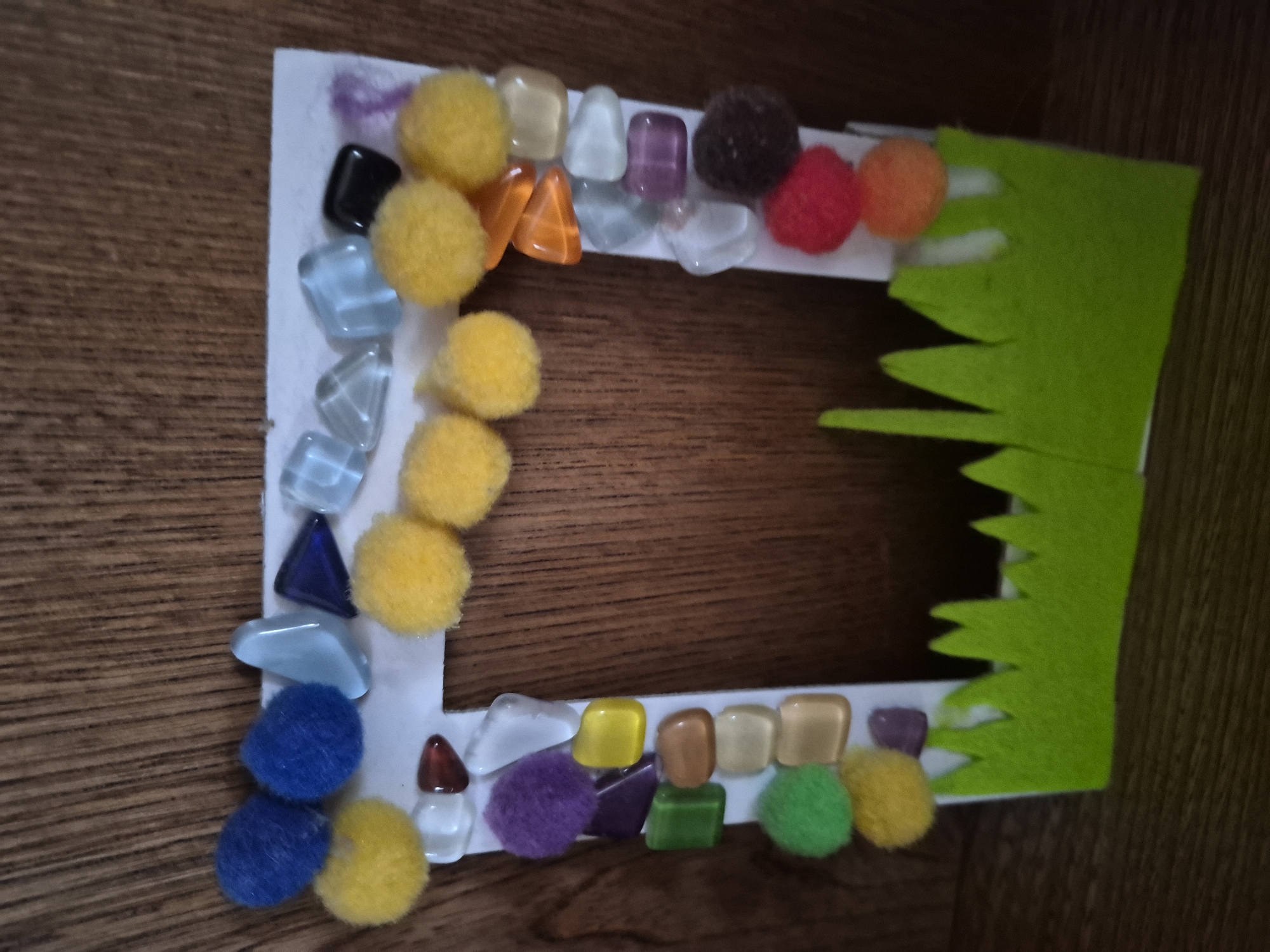 An ornate, handmade picture frame is displayed on a wooden surface. The frame is decorated with colorful pom-poms and assorted glass beads in various shapes and colors, including blue, yellow, purple, green, orange, and red. The bottom of the frame is adorned with a green felt cutout resembling grass. The background is a vertical wooden plank with a visible grain. The lighting appears to be natural, casting soft shadows. No people are present in the image.