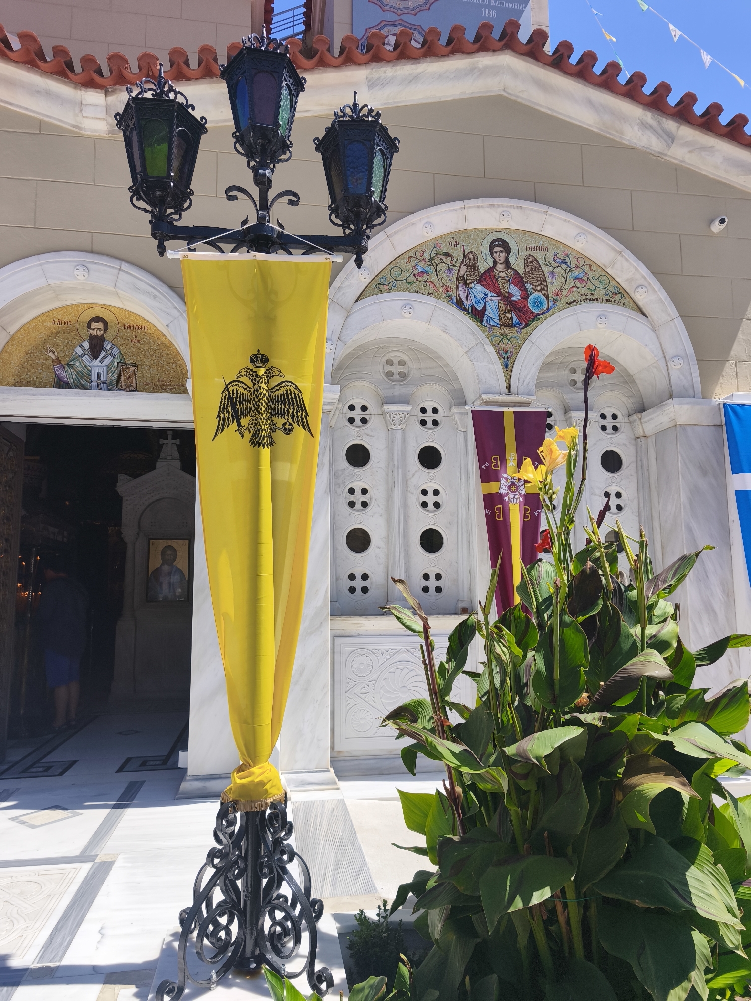 The image shows the entrance to a church in Prokopi, Greece. A tall, ornate black lamppost with three colored glass lanterns stands in front of the entrance. A yellow banner with a double-headed eagle crest is draped over the lamppost. To the right of the entrance, a potted plant with large green leaves and red and yellow flowers is visible. The church facade is made of white marble and decorated with mosaics above the arched entrance. The ground in front of the church is paved with white and grey marble tiles. A portion of a blue flag with a white cross is visible on the far right of the image. The overall scene suggests a religious ceremony or event is taking place.
