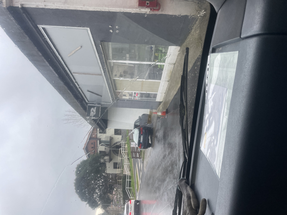 The image depicts a street scene in the city of Zestoa, Spain, under overcast, wet conditions. The vantage point is from inside a vehicle, looking through a severely cracked windshield covered with numerous water droplets. The dashboard is visible in the foreground, featuring a gray work glove on the left and a document in a clear plastic sleeve on the right.
In the midground, a wet asphalt road leads into the distance. A black BMW sedan with license plate "7177 BPW" is parked partially on the curb to the right of the road. Further left, a white SUV is partially visible, moving away from the viewer. An orange traffic cone stands on the sidewalk near the front of the black BMW.
To the right, a dark gray building with a white base and a flat roof dominates the midground. It features a sign with a stylized "22" or similar logo. The upper section of the building contains large, rectangular windows, appearing to be boarded or covered from the inside with visible wooden supports. The lower section consists of multiple retail-style display windows, reflecting elements of the street scene.
In the background, beyond the road and to the left, green foliage and trees are visible, along with traditional residential buildings featuring pitched roofs and balconies, suggesting a hilly terrain. The sky is uniformly gray and overcast. No individuals are visible in the scene.