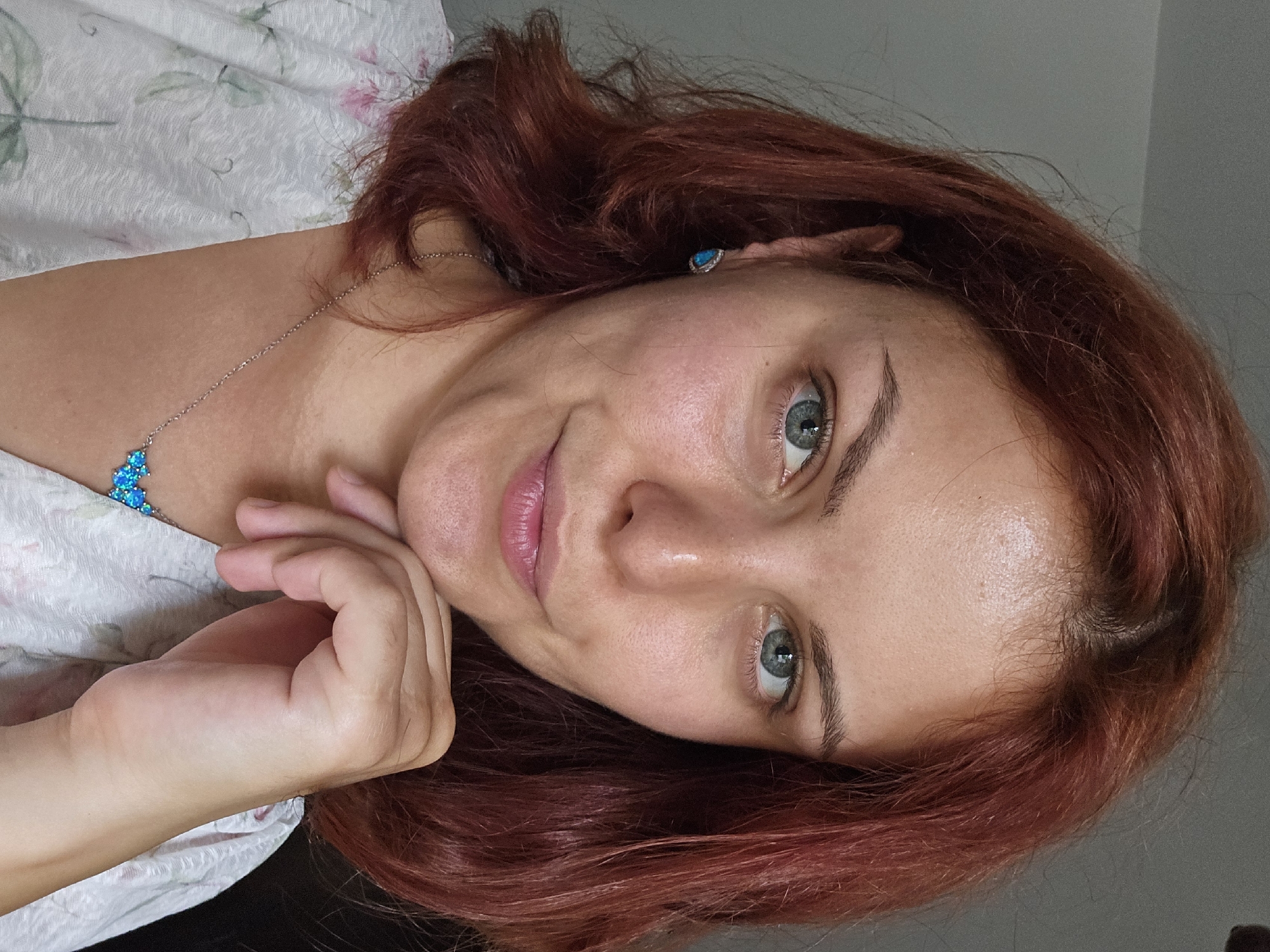 This is a close-up, rotated portrait of a woman with auburn hair. She is looking towards the viewer with a gentle smile. Her left hand is resting on her cheek, and her head is tilted. She is wearing a white blouse with a floral pattern and a silver necklace with blue pendants. Her hair is shoulder-length and appears slightly messy, framing her face. The background is a plain grey wall, and the lighting suggests an indoor setting, possibly during the daytime. Her expression is relaxed and approachable. There is no visible text in the image.