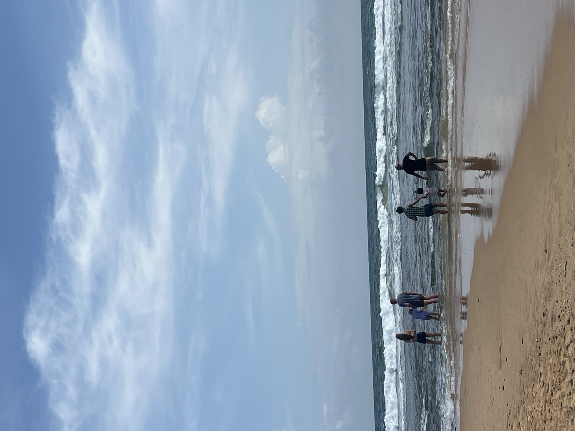 This is a full-frame, eye-level shot taken on a sandy beach in Hikkaduwa, Sri Lanka. The scene captures a group of six individuals, including adults and children, wading in the ocean. The water is a dark blue-green, with a prominent line of white-capped waves breaking closer to the shore and smaller waves further out. The beach is composed of light brown sand, and a reflection of the people and the sky is visible on the wet sand near the water's edge. The sky above is a bright blue, with scattered white, wispy clouds. The overall atmosphere is bright and summery, with the ocean and sky dominating the upper two-thirds of the frame and the sandy beach occupying the foreground.
