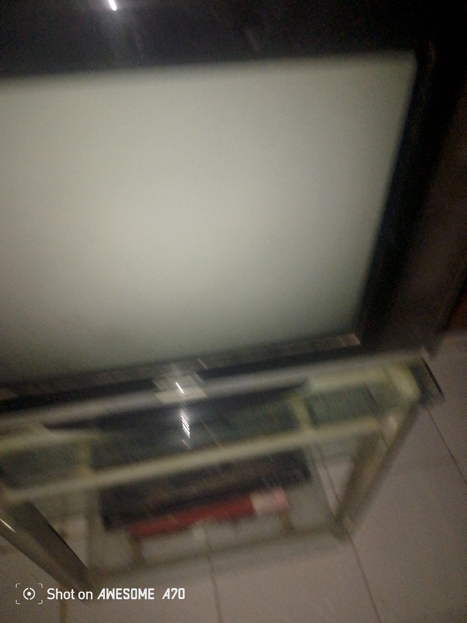 The image shows a television set on a glass stand. The television screen is blank and off-white, suggesting it is not powered on or is displaying a static image. The television set appears to be older, possibly a cathode ray tube (CRT) television, given its thickness. The glass stand has multiple shelves and metal supports, holding some unidentifiable items on the lower shelves. The lighting in the scene is dim, with a bright light source visible at the top of the image, casting a glare on the television screen. A watermark in the bottom left corner reads "Shot on AWESOME A70". The floor appears to be tiled. The overall setting is indoors, likely a living room or entertainment area. The context of Jalingo, Nigeria is not visually evident from the image itself.