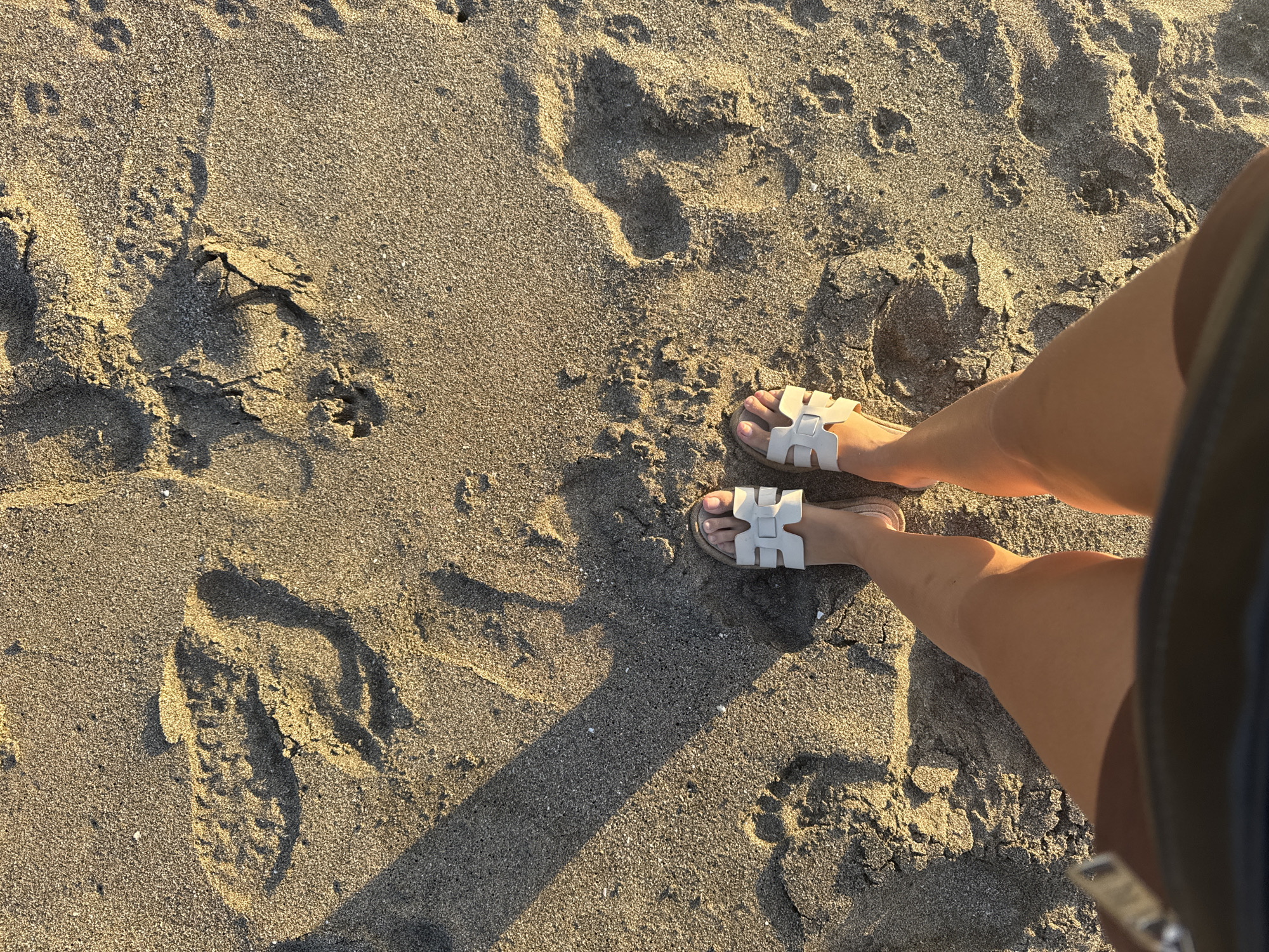 From an elevated perspective, the image captures a person's legs and feet standing on a sandy beach. The person is wearing white sandals. The sand is light brown and coarse, marked with numerous footprints. Shadows cast by the person and their surroundings indicate the presence of a light source, likely the sun. The setting is presumed to be a beach, potentially in North Kuta, Indonesia.