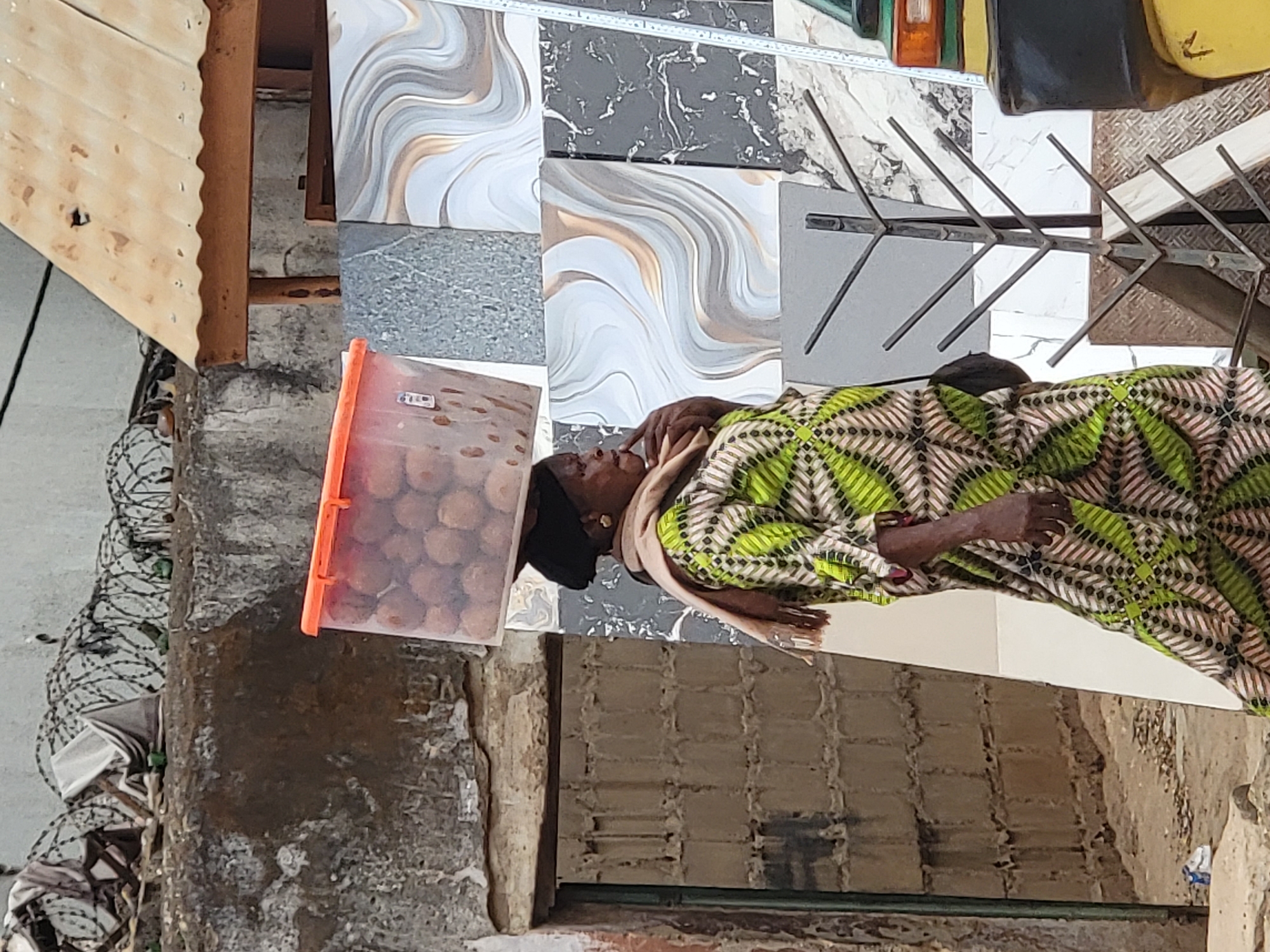 In Maiburiji, Nigeria, an older woman with dark skin stands in an urban environment. She wears a head covering, a tan scarf, and a green and brown patterned dress. In front of her is a clear orange plastic container filled with round objects, likely food items for sale. She stands near a display of large ceramic tiles with swirling gray, white, and brown designs, as well as solid gray tiles. Behind her is a weathered wall with exposed brickwork and deteriorating plaster. A metallic rack structure stands nearby. The scene appears to be an outdoor marketplace or roadside vending spot.