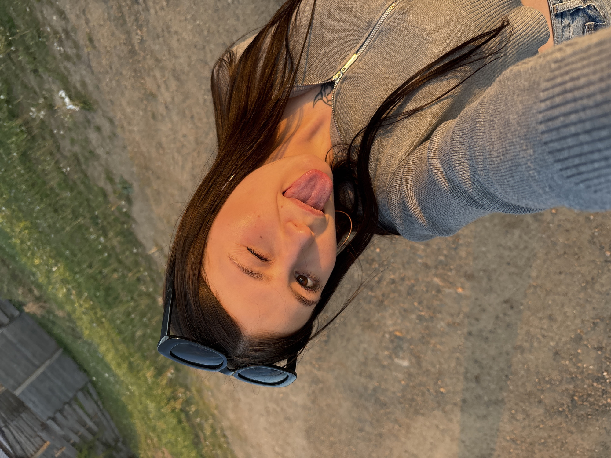 A woman with long, dark hair is taking a selfie, sticking her tongue out and winking. She is wearing a gray zip-up jacket and jeans. She has sunglasses perched on her head and hoop earrings. There is a tattoo visible on her neck. The background appears to be a dirt or gravel path. The location in the city of Фоки, Russia is not discernible from the image.