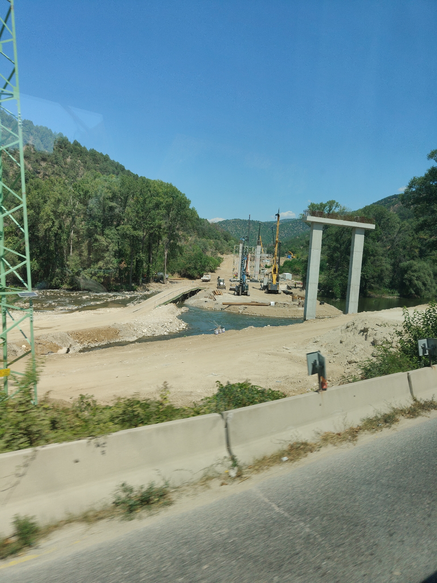 The image depicts a construction site for a bridge over a river, likely located near Kresna, Bulgaria. A large green metal structure, possibly part of an electrical pylon or crane, is on the left side of the frame. The river flows through a valley with tree-covered hills on either side. In the middle ground, construction equipment, including cranes and excavators, is visible. A partially constructed concrete structure, resembling a bridge support, stands prominently in the right-middle section of the image. The ground is a mix of dirt and gravel, and a temporary bridge is present, spanning a section of the river. The sky is clear blue, indicating a sunny day. The foreground shows a concrete barrier and a road, suggesting the image was taken from a vehicle.
