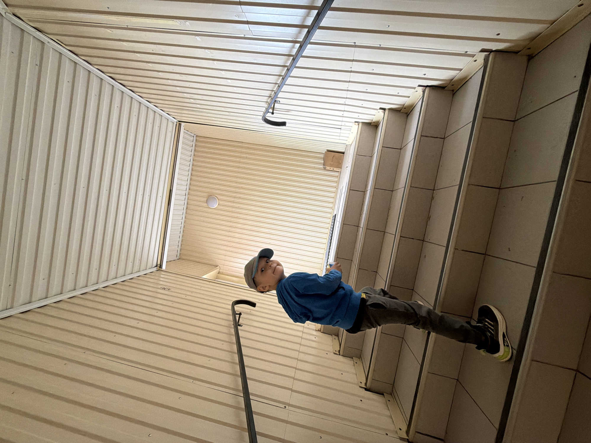 A low-angle shot in what appears to be an industrial or commercial location in Chaikovsky, Russia. The walls and ceiling are constructed of corrugated metal panels, indicating a warehouse or storage facility. A boy, wearing a blue long-sleeve shirt, gray pants, a baseball cap, and black sneakers with neon green accents, is seen standing near a staircase on the right side of the frame. A handrail runs along the left wall. The camera is tilted, creating a disorienting perspective. The boy is looking up and possibly holding an object in his hand, though the detail is obscured.