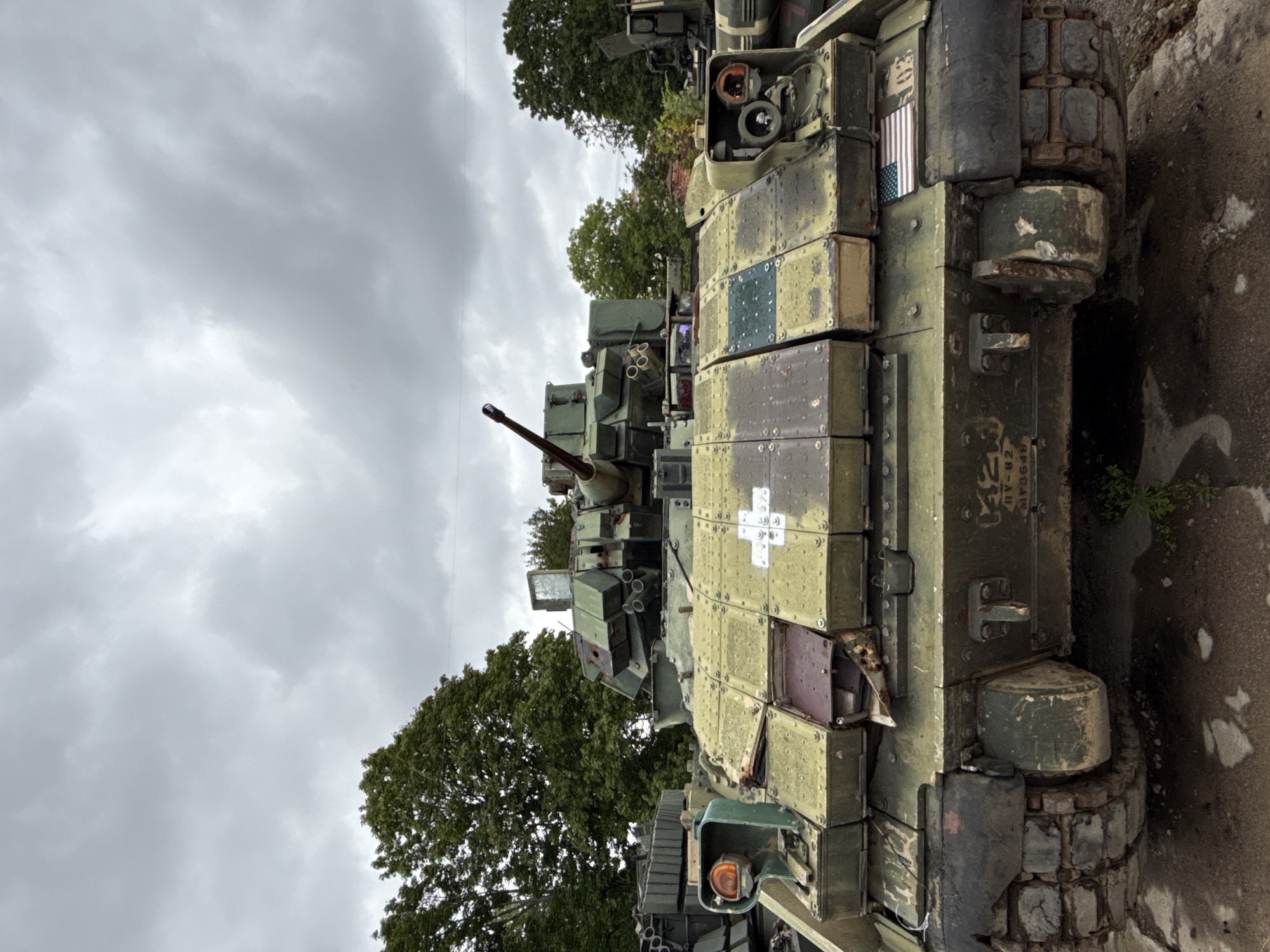 The image shows the front view of a damaged military tank parked outdoors. The tank is predominantly olive green with patches of rust and wear. It has visible tracks, a turret with a cannon, and various hatches and armor plating. A white cross is painted on the front left side of the armor. An American flag decal is visible on the front right side. The tank is positioned in what appears to be an outdoor storage or display area, with trees and a cloudy sky in the background. There are no people present in the image. The tank is located in the city of Kronstadt, Russia.