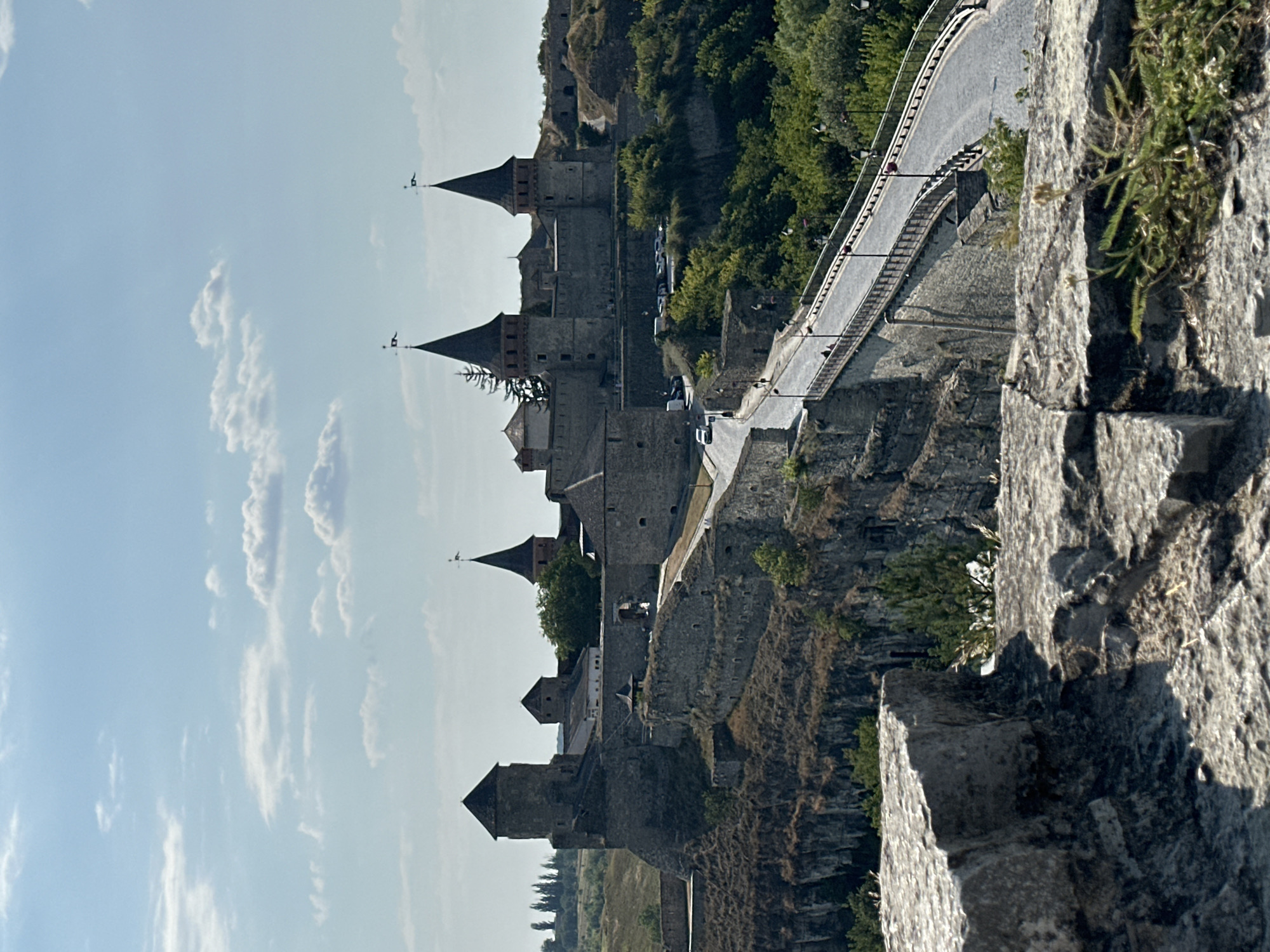 The image depicts a panoramic view of the Kamianets-Podilskyi Castle, a historic fortress located in Kamianets-Podilskyi, Ukraine. The castle, constructed from grey stone, is perched atop a steep rocky cliff. Several towers with pointed roofs, some adorned with flags, rise prominently from the structure. A paved road, lined with lampposts, curves across a bridge and leads towards the castle's entrance. The surrounding landscape is characterized by lush green vegetation and a bright, partly cloudy sky. In the foreground, rough-hewn stone blocks suggest the viewer is positioned on another part of the castle's fortifications. Two white cars are visible on the road leading to the castle.