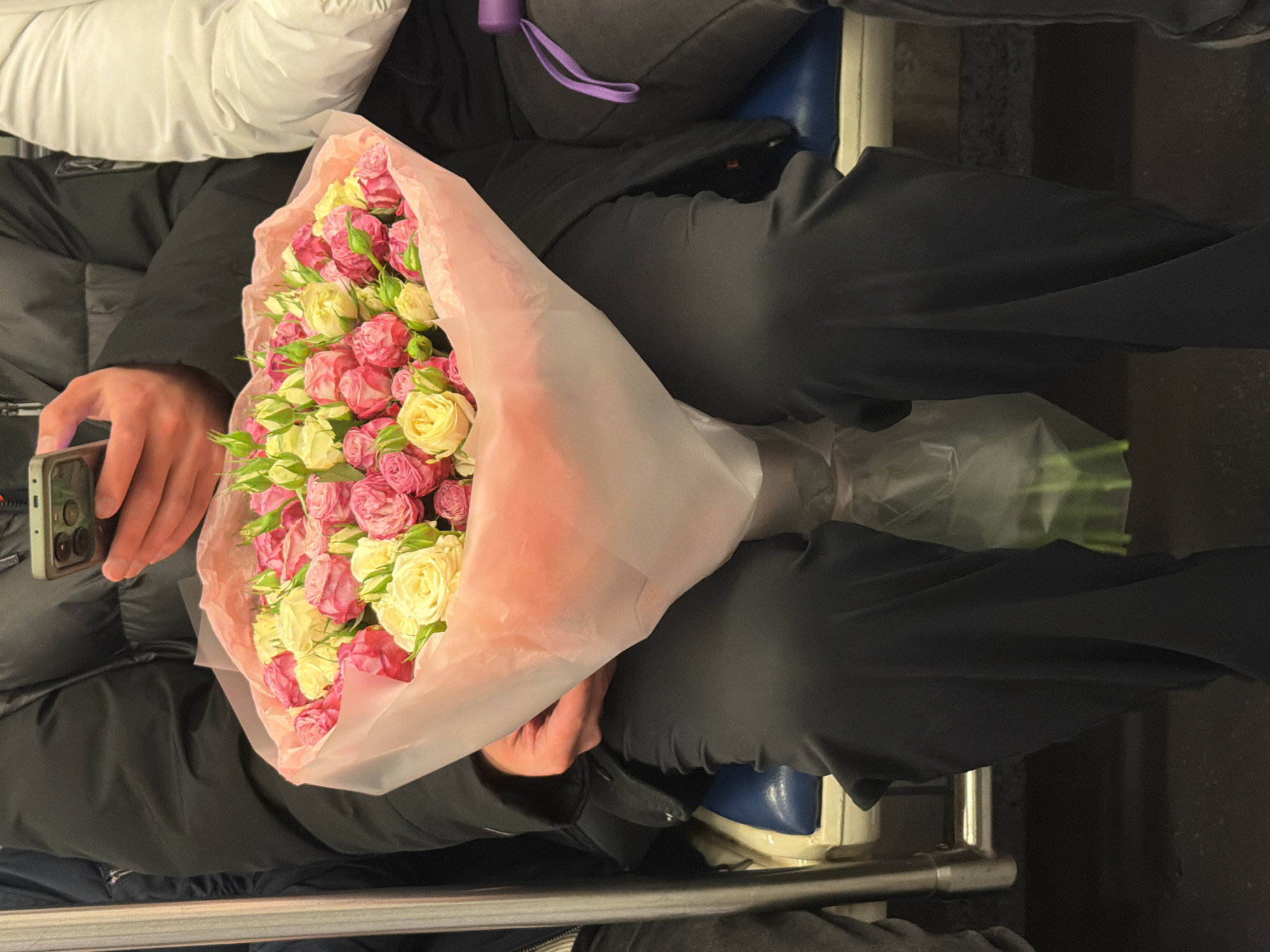 This image, set in Moscow, Russia, captures a close-up view of two individuals seated, likely within a public transport vehicle such as a subway or train car. The photo is taken from a slightly elevated perspective, looking down into the seated area.
In the foreground, a person is holding a large, vibrant bouquet of small roses. The roses are a mix of bright pink and creamy yellow hues, tightly packed, and wrapped in translucent light pink floral paper. The person is wearing a black puffer jacket and dark trousers. Their left hand is visible, holding a smartphone with a light-colored case featuring multiple camera lenses. Their right hand supports the bouquet.
To the right and slightly behind the first person, another individual is partially visible. They are also wearing dark trousers. A white padded surface, possibly a headrest or part of the seat, is visible above them. A purple strap, perhaps from a bag or water bottle, hangs near their torso. Their legs are visible, and they appear to be wearing blue shoes with white soles. Beneath their legs, a metal handrail or safety bar runs horizontally across the frame.
In the bottom right corner of the image, another clear plastic-wrapped bundle, possibly containing more flower stems or another bouquet, can be seen on the floor or seat. The seating appears to be blue, consistent with public transport. The overall setting is enclosed, suggesting an indoor environment. No specific text is visible within the frame. The presence of the bouquet suggests a special occasion or gift.