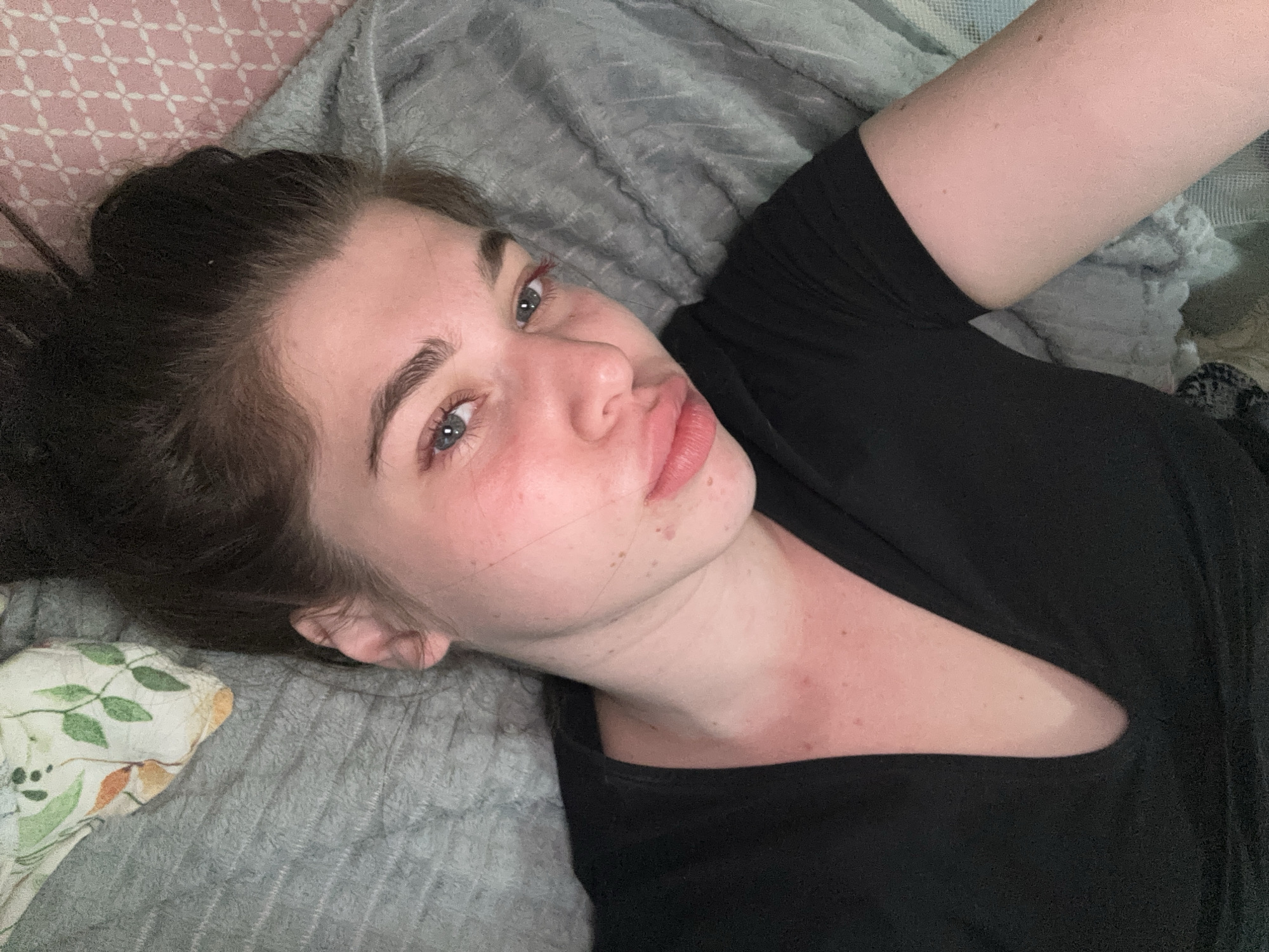 A woman is lying on a gray blanket. She has dark hair pulled back, light skin, and blue eyes. She is wearing a black shirt. The background includes a pink patterned surface and a pillow with a floral design.
The location is not apparent in this image. The query suggests a city in Мещёры, Russia, but there is no supporting evidence in the image to confirm this.