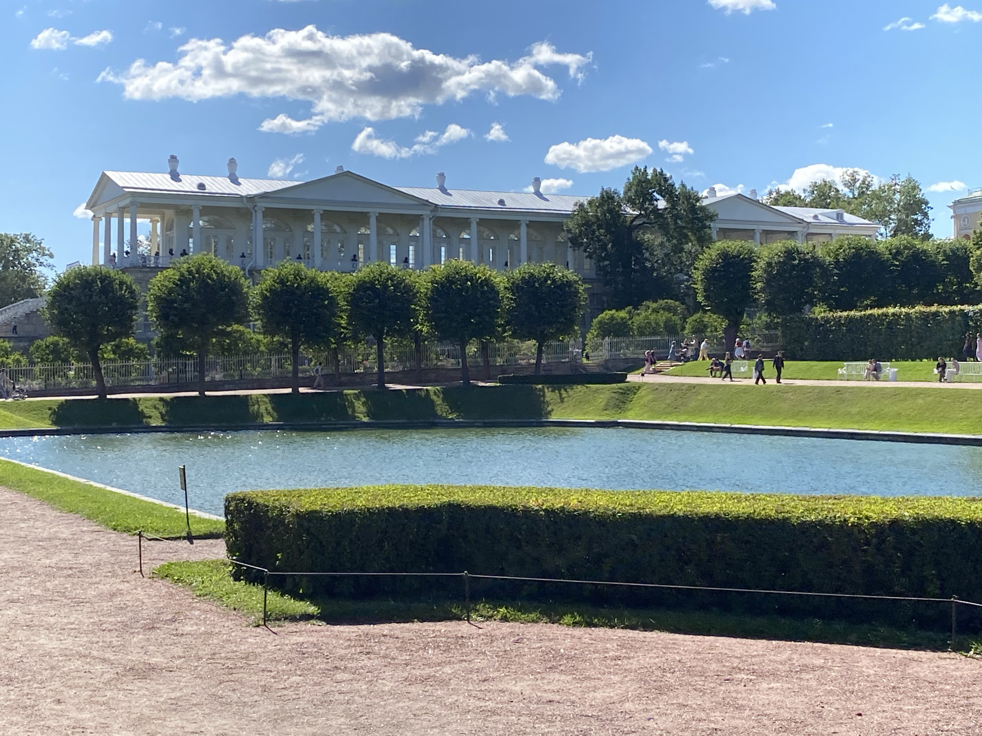 A bright, sunny day is depicted in this outdoor shot of a classical European-style palace and its grounds. In the foreground, a path made of gravel leads the eye toward a tranquil blue pond. A neatly trimmed hedge borders the pond, separating it from the gravel path. Beyond the pond, a lush green lawn stretches out, dotted with several small, dark green trees with rounded tops.
In the mid-ground, a grand, white palace with a long colonnade of tall, slender columns stands proudly. The palace has a light-colored roof with two prominent chimneys. Several people are visible on the terrace in front of the palace, and a few more are scattered across the lawn.
The background shows more trees and greenery, suggesting a large estate or park. The sky is a vibrant blue, punctuated by fluffy white clouds, indicating pleasant weather. The overall atmosphere is serene and picturesque, hinting at a leisurely day spent exploring the historical site. The architecture and landscaping suggest a location with significant historical and cultural value.
