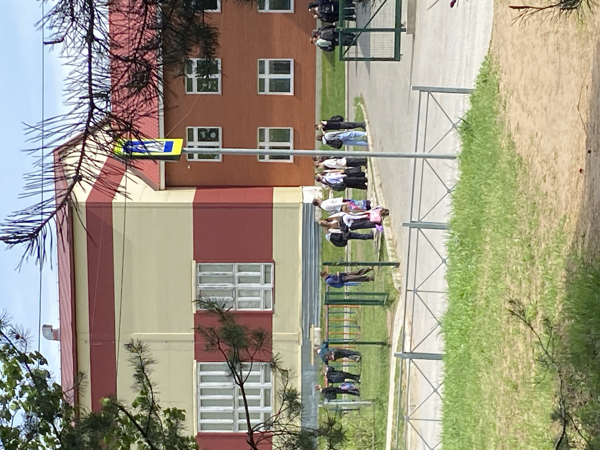 Here's a description of the image:
The image shows a group of people, likely students, standing outside a building. They are standing in a line, possibly waiting for something. The building is two-toned, with a red and yellow exterior. There are several windows visible on the building. A yellow and blue pedestrian crossing sign is seen attached to the building. The surrounding area is green, with trees and grass. Based on the available information, the location is in Noviy, Russia.