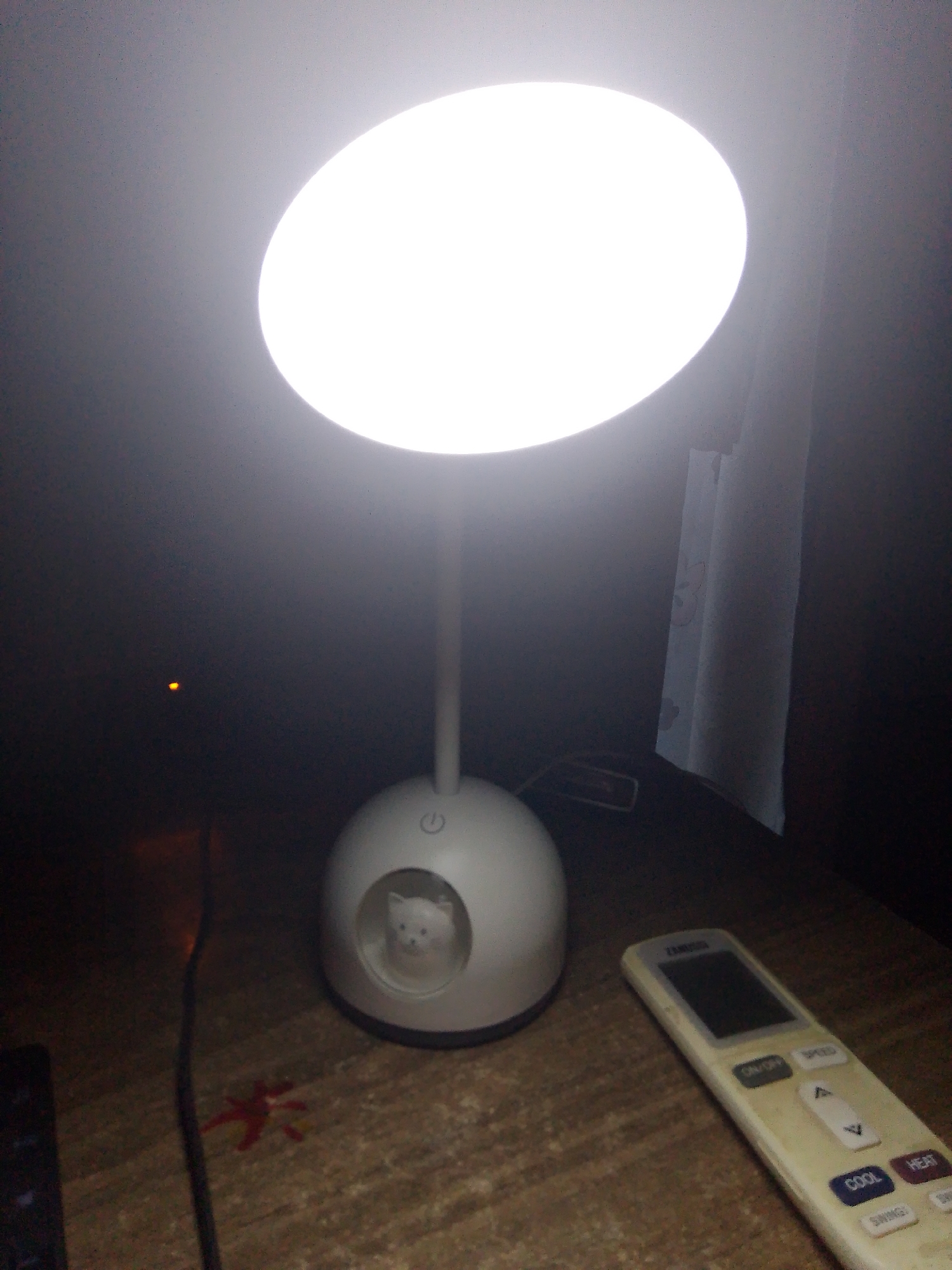 The image depicts a desk lamp with a circular, illuminated top casting a bright light. The base of the lamp is round and white, with a small, clear circular cutout containing a white cat figurine. A power button icon is visible on the front of the base. A cream-colored remote control, likely for an air conditioner, lies on the wooden surface in front of the lamp. To the left of the lamp, a dark object, possibly a keyboard, is partially visible. A red, star-shaped object is on the wooden surface to the left of the lamp's base. In the background, a curtain is visible to the right of the lamp. A small, orange light source is present in the dark background to the left of the lamp. The image is taken in a dimly lit room. The location of this scene within the city of Batumi, Georgia, cannot be determined from the provided image.