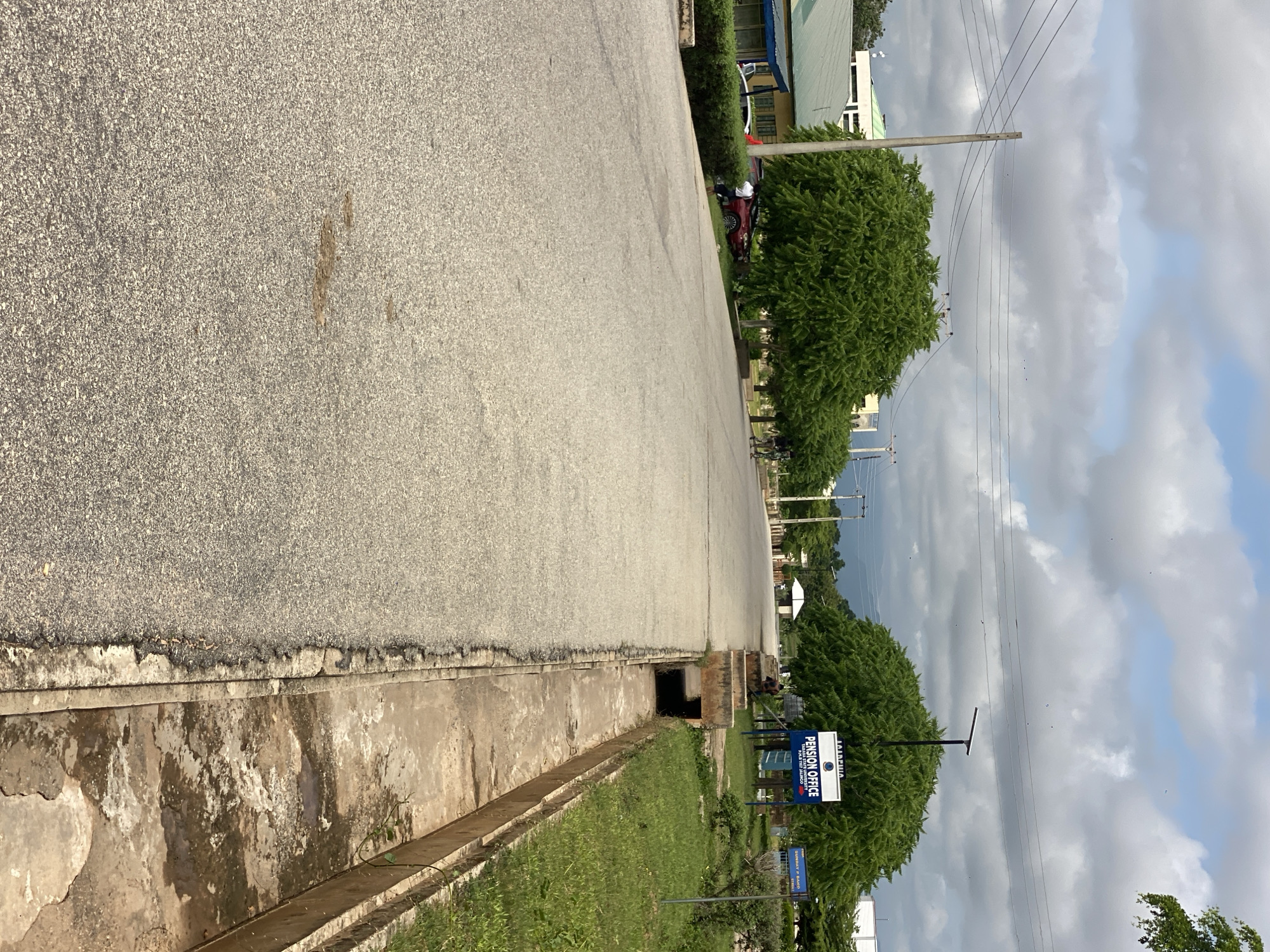 The image depicts a street scene in Jalingo, Nigeria, on a cloudy day. A paved road occupies the foreground and midground, with a concrete drainage ditch running along its right side. Lush green trees line the sides of the road, some with dense canopies. In the background, buildings and utility poles are visible. Several people are present, including a person on a scooter in the distance on the right side of the road. Two vehicles are parked on the left side of the road. A blue sign with white lettering, bearing the text "PENSION OFFICE" and "TARABA STATE UNIVERSITY," is prominently displayed amidst the trees. The sky is filled with scattered clouds, with some darker formations suggesting potential rain. The overall impression is of a well-maintained urban environment with significant vegetation.