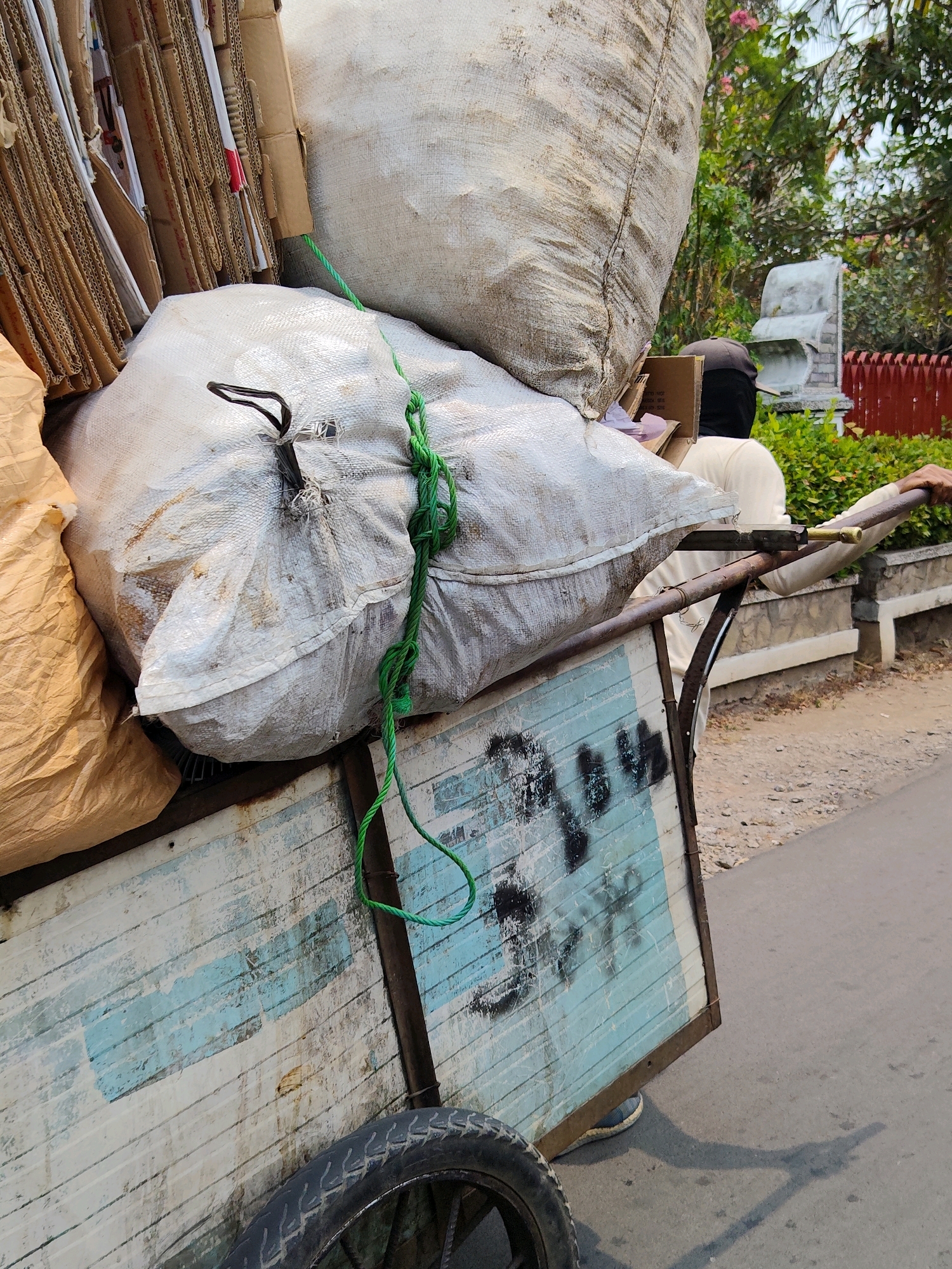 A person is pushing a cart laden with large sacks and cardboard boxes along a paved road in Purwakarta, Indonesia. The cart has a weathered blue and white painted side with black graffiti. The person, partially obscured, wears a light-colored shirt and a dark cap. The sacks are white and tan, and several flattened cardboard boxes are stacked on the cart. In the background, there is foliage, a red fence, and a stone structure. The sunlight casts shadows on the road.