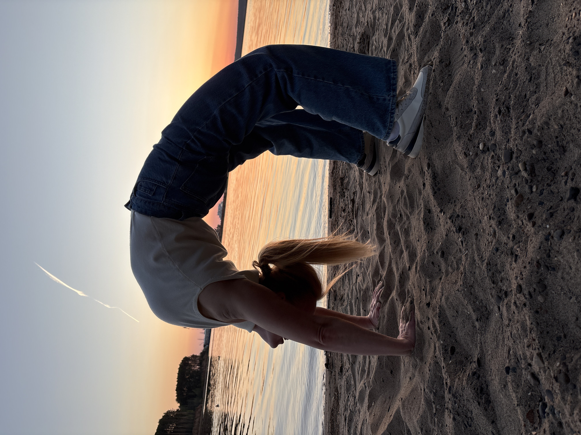 A woman performs a backbend on a sandy beach at sunset. She wears a white t-shirt and loose-fitting denim pants with white sneakers. Her hair is blonde and pulled back in a ponytail. The background shows a body of water reflecting the colors of the setting sun, and the shoreline is visible in the distance. The location is likely near the city of Chaikovsky, Russia, as it is near the shoreline. The sand is dark with pebbles and footprints, indicating human activity in the area.