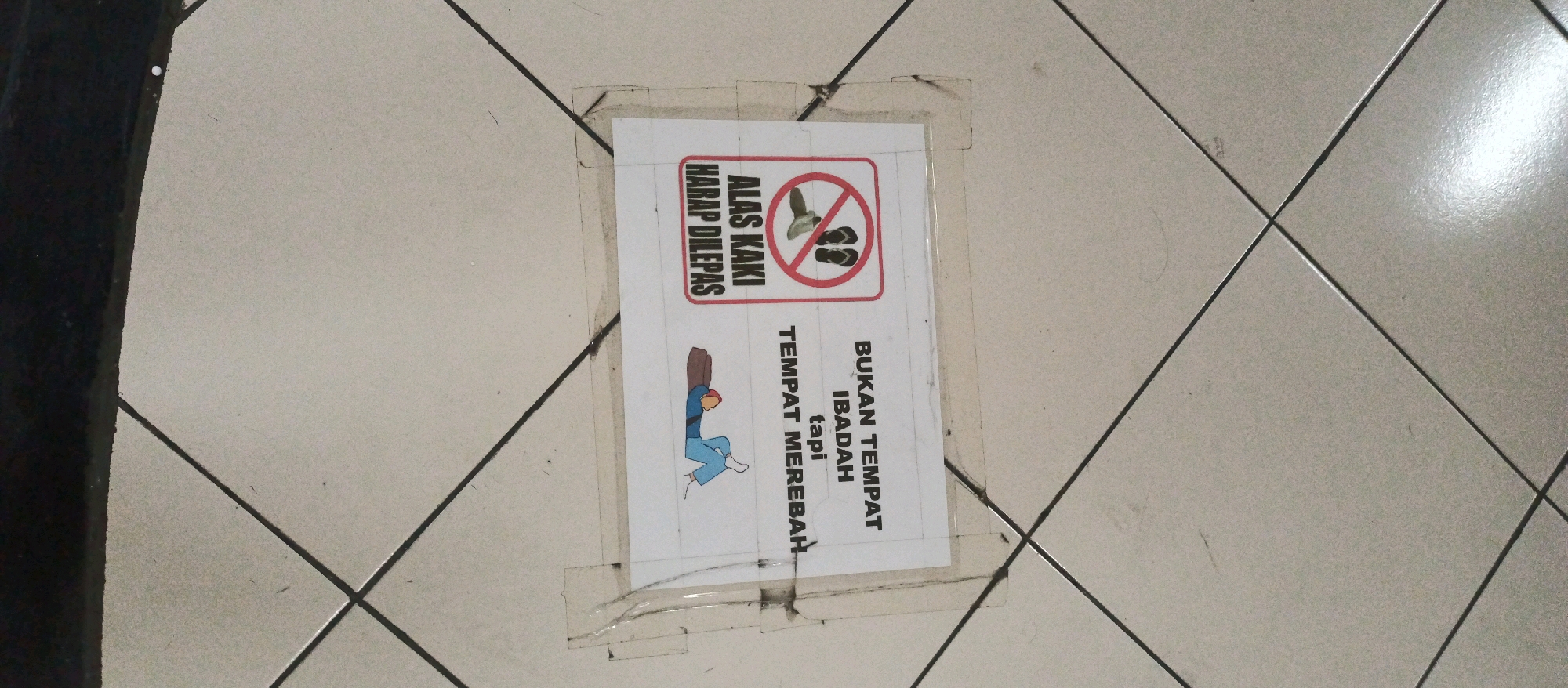 A sign taped to a tiled floor reads "ALAS KAKI HARAP DILEPAS" which translates to "Shoes Please Remove". Below this, a pictogram depicts a person removing their footwear. The sign also states "BUKAN TEMPAT IBADAH tapi TEMPAT MEREBAH", meaning "Not a place of worship but a place to lie down". The floor tiles are light grey and arranged in a diamond pattern. The sign appears to be laminated and secured with clear tape. To the left of the sign, a dark vertical object is visible, possibly a wall or pillar. The lighting suggests an indoor environment.