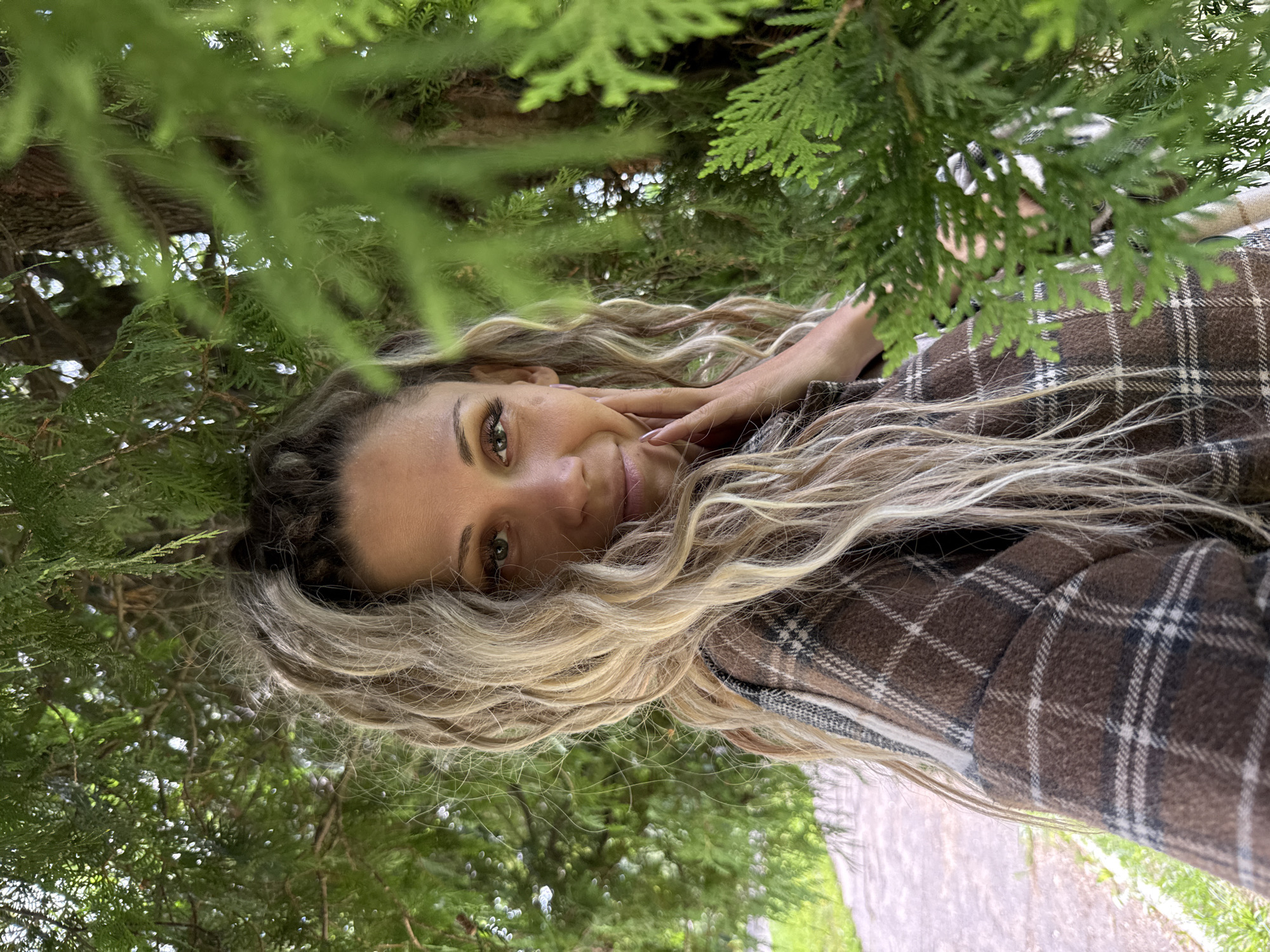 A woman with long, wavy blonde hair, wearing a brown plaid jacket, is pictured in an outdoor setting. She is positioned behind some lush green foliage, with some of the leaves and branches obscuring parts of her face and body. The woman is looking towards the camera with a slight smile. Her right hand is raised to her cheek. In the background, a paved path and green grass are visible, suggesting a park or natural area. The overall lighting suggests it is daytime. The provided location is the city of Rusanovo, Russia.