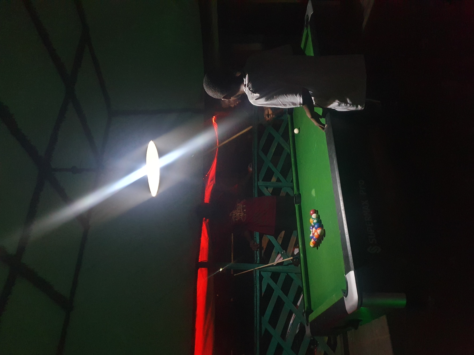 In Lokoja, Nigeria, a pool hall features a green pool table with a rack of colorful balls at one end and a single white ball near the other. A young boy, viewed from behind and wearing a light gray shirt, stands facing the table, his hand resting on its edge. In the background, a person in a red shirt stands near a pool cue leaning against a railing. The scene is dimly lit by a bright overhead light, casting strong beams and shadows across the room, illuminating the ceiling structure and a section of the wall.
