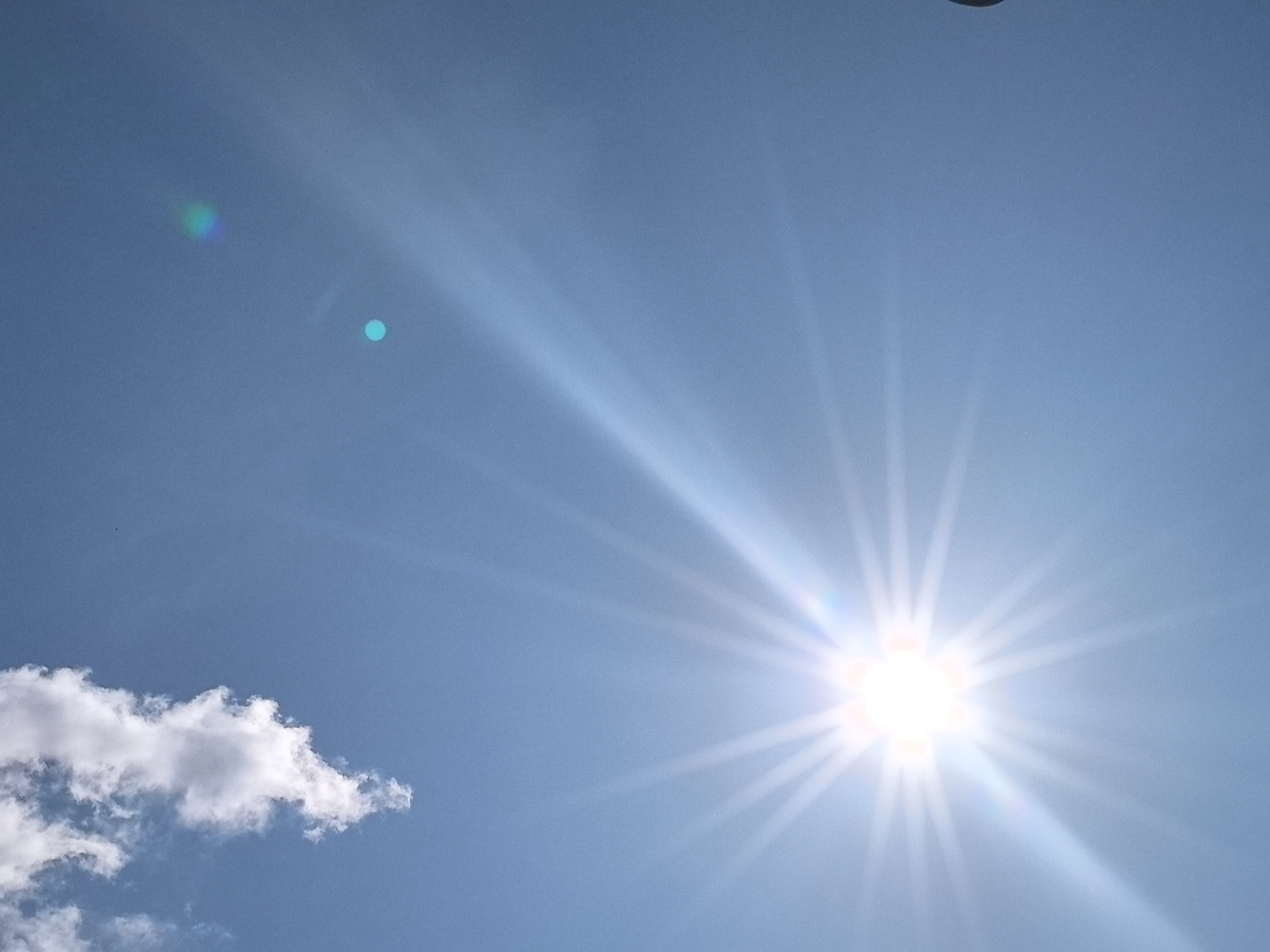 The image depicts a bright sunny day with a clear blue sky. The sun is prominently visible in the center-right of the frame, emitting strong rays of light that create a lens flare effect. A few wispy clouds are scattered across the sky, with one larger cloud formation in the bottom left corner. There are also two small, indistinct circular light anomalies, possibly lens artifacts or distant objects, visible in the upper left portion of the sky. The scene appears to be an outdoor environment, likely in an open area, given the unobstructed view of the sky and the presence of clouds. No people or specific man-made structures are identifiable in the image, suggesting a natural or semi-rural setting. The location is identified as Опенок-1 Снт, Russia.
