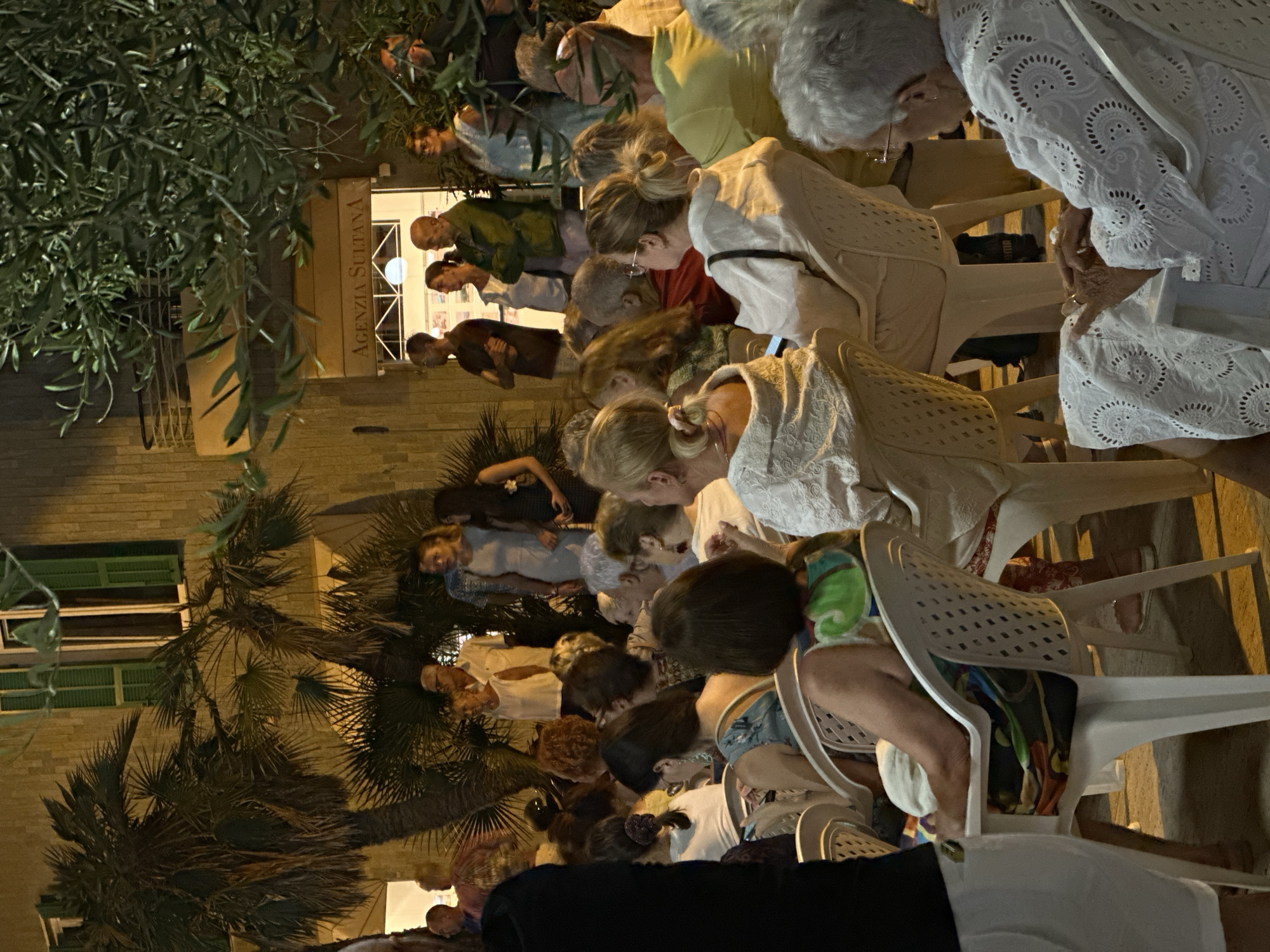In Ospedaletti, Italy, an outdoor evening gathering is underway, with a crowd of approximately 30-40 individuals seated on white plastic chairs arranged in rows. The event appears to be a performance or presentation, as many attendees are facing forward, towards an unseen focal point. The background features a building with a sign reading "AGENZIA SULTANA," indicating a commercial establishment, possibly a real estate agency. Palm trees and olive branches frame the scene, suggesting a Mediterranean setting. The lighting is warm and artificial, characteristic of evening events. The attire of the attendees varies, from casual to slightly more formal, with some individuals wearing light-colored clothing. The mood seems relaxed and engaged, with people observing, listening, and interacting amongst themselves.