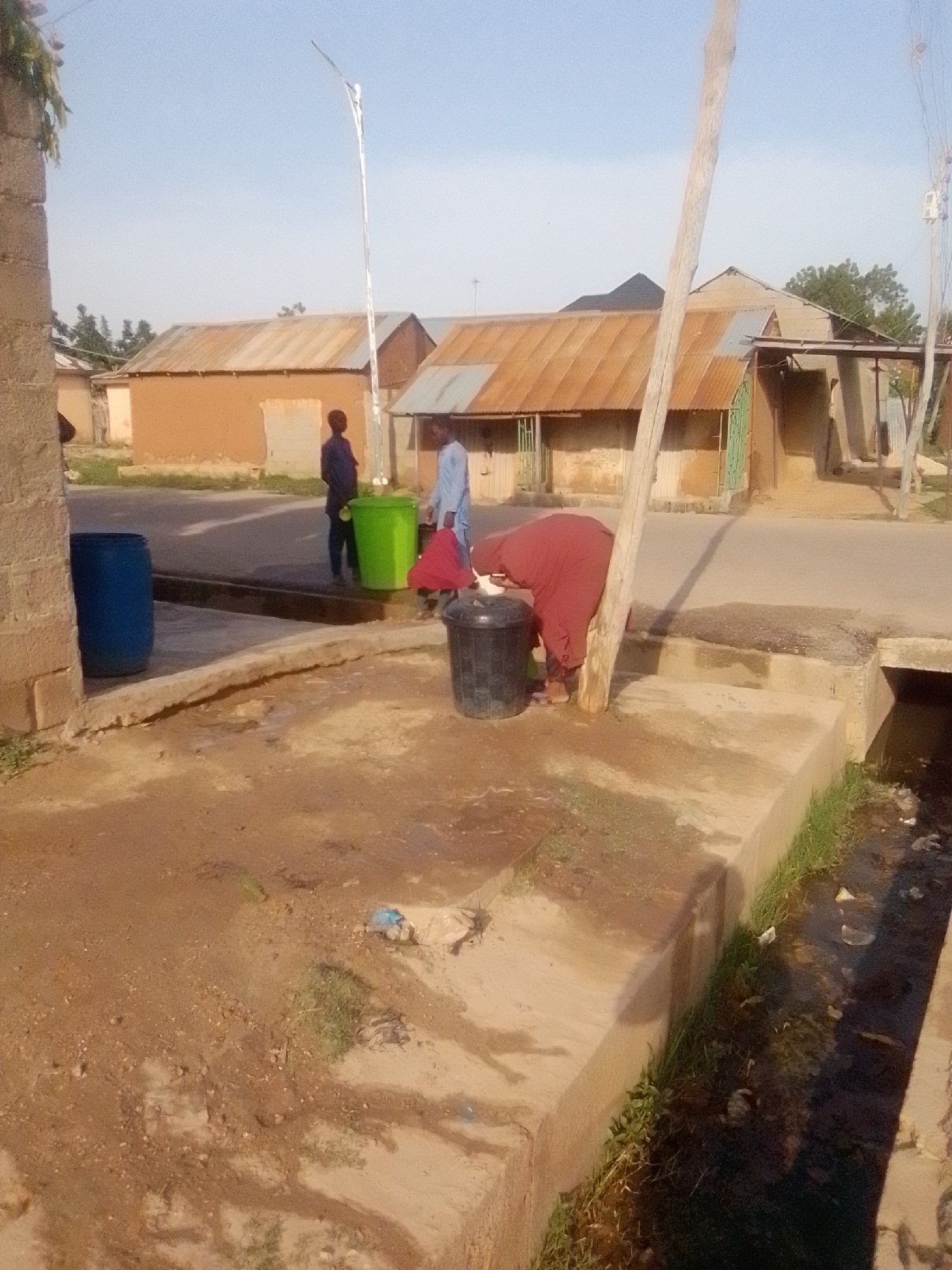 The image depicts an outdoor scene in Gombe, Nigeria, characterized by a dusty, unpaved ground and a partial view of residential structures with corrugated metal roofs. In the foreground, a woman in a maroon garment is seen bending over a black trash bin, appearing to be cleaning or disposing of something. Adjacent to her, a large blue barrel is positioned near a concrete curb. To the left, a partially visible brick structure suggests it is part of a building. Further back, two young males are present: one in a dark blue shirt stands near a bright green trash bin, and another in a light blue shirt is seen walking away from the green bin. The background reveals the distinct architecture of modest dwellings. A utility pole with electrical wires is also visible. The overall environment suggests a common area within a residential neighborhood.