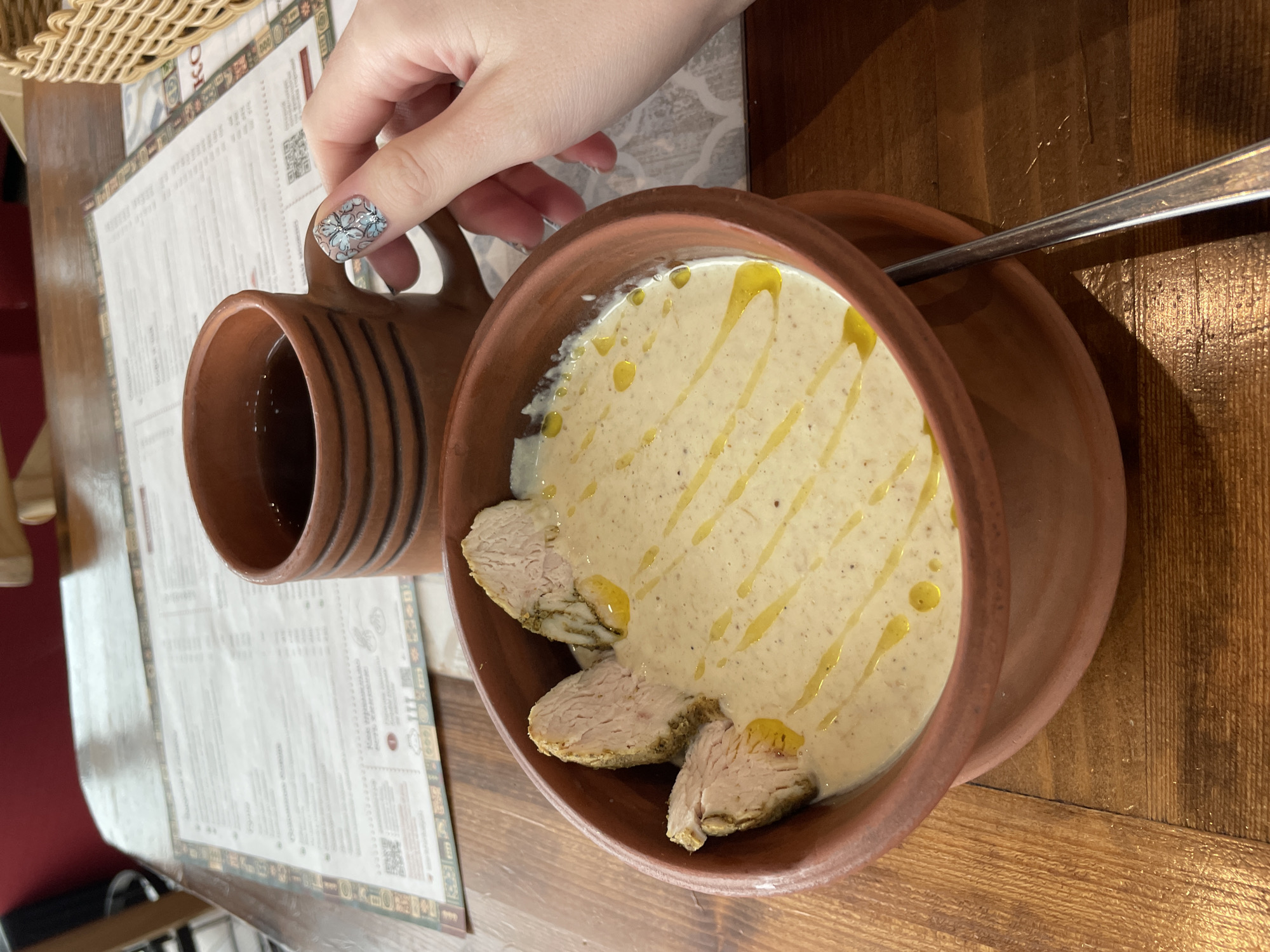 A close-up, medium-shot photograph displays a meal served on a wooden table. At the center, a brown ceramic bowl holds a creamy soup drizzled with oil. The soup is garnished with slices of meat arranged around its edge. To the left, a matching brown ceramic mug contains a dark liquid, possibly tea or coffee, and is partially held by a hand with patterned nail art. In the background, a printed menu with Cyrillic text is visible, along with a glimpse of a woven basket. A portion of a spoon rests on the right side of the soup bowl. The location is suggested to be in Izhevsk, Russia.