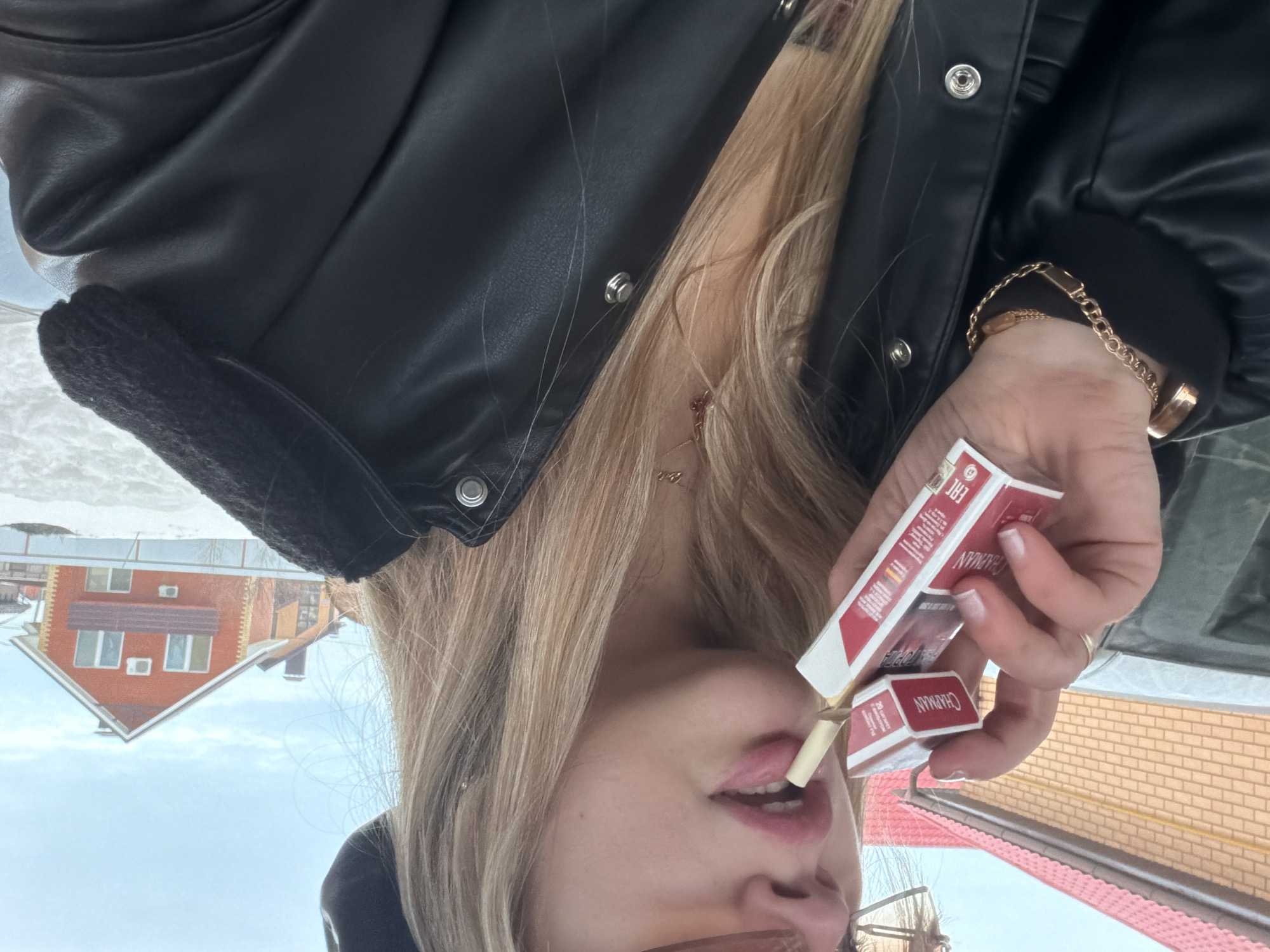 A medium close-up shot captures an individual outdoors in a residential setting in Большая Шильна, Russia. The subject, a person with long blonde hair, is partially visible from the chest up, oriented slightly to the right of the frame's center. They wear a black faux leather jacket with metallic snap fasteners, a dark beret-like hat on their head, and amber-tinted sunglasses pushed up into their hair. A gold-colored chain necklace with a small pendant and a gold-colored chain bracelet on the left wrist are visible. The subject's lips, coated with pink lipstick, are slightly parted, holding the filter end of a light-colored cigarette. Their left hand holds two red and white cigarette packs, both labeled "CHAPMAN" with Russian text and an EAC conformity mark. One cigarette is partially extracted from a pack.
The background shows a snowy ground. A prominent two-story house with an orange-red facade, a brown gabled roof, and multiple windows with white frames is situated in the mid-ground. Two white outdoor air conditioning units are mounted on its front wall. A dark-colored vehicle is partially visible behind the subject to the left. Another brown brick building with a red roof is partially visible further left. The sky above is light blue with scattered white clouds.