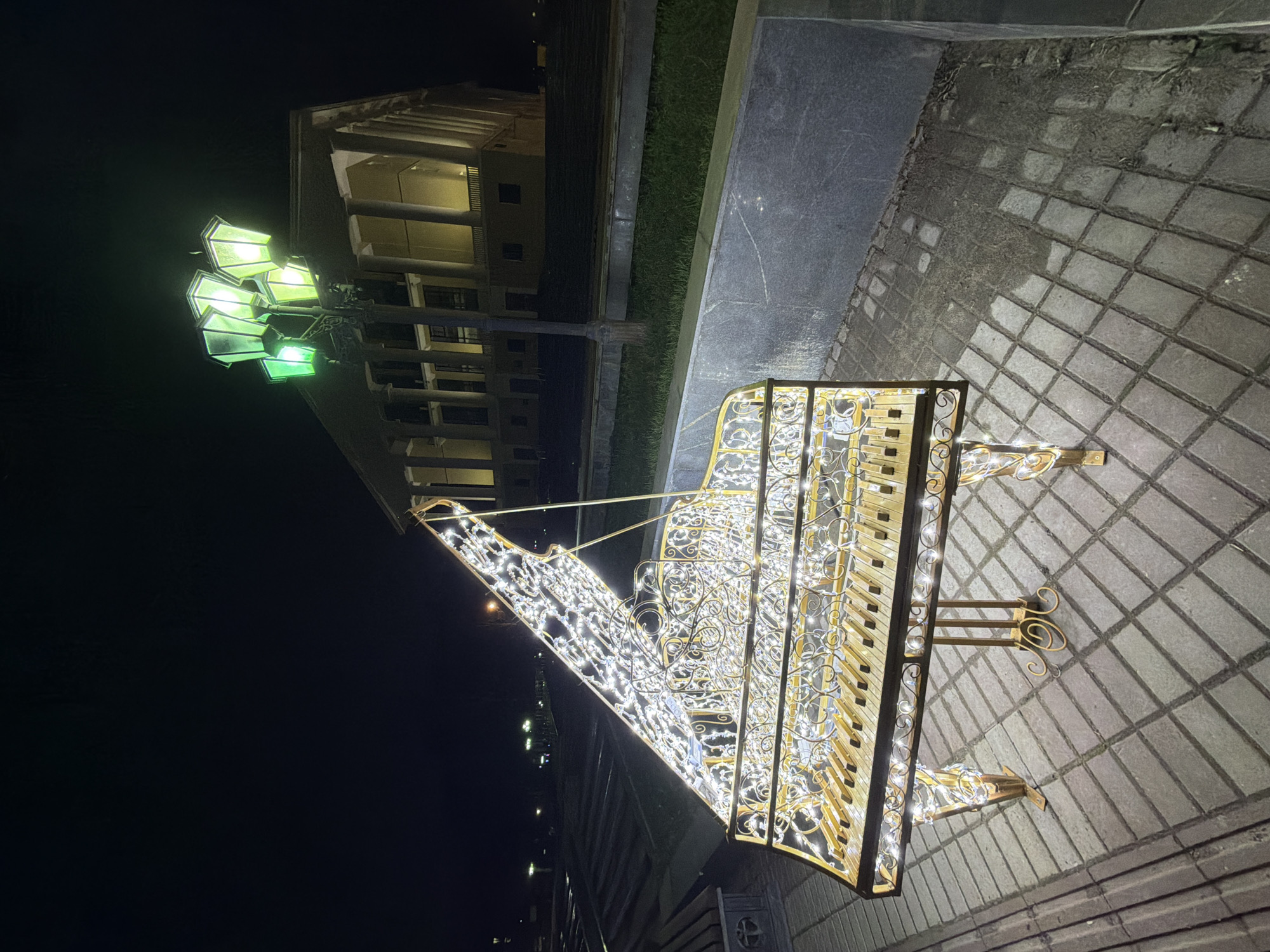 The image shows an illuminated ornamental piano sculpture located in Votkinsk, Russia, at night. The sculpture sits on a paved area, with a building featuring lit colonnades in the background, and a green lamppost illuminating the scene. The piano is decorated with numerous small white lights. The surrounding environment is dark, with only the lights from the sculpture and lamppost providing illumination.