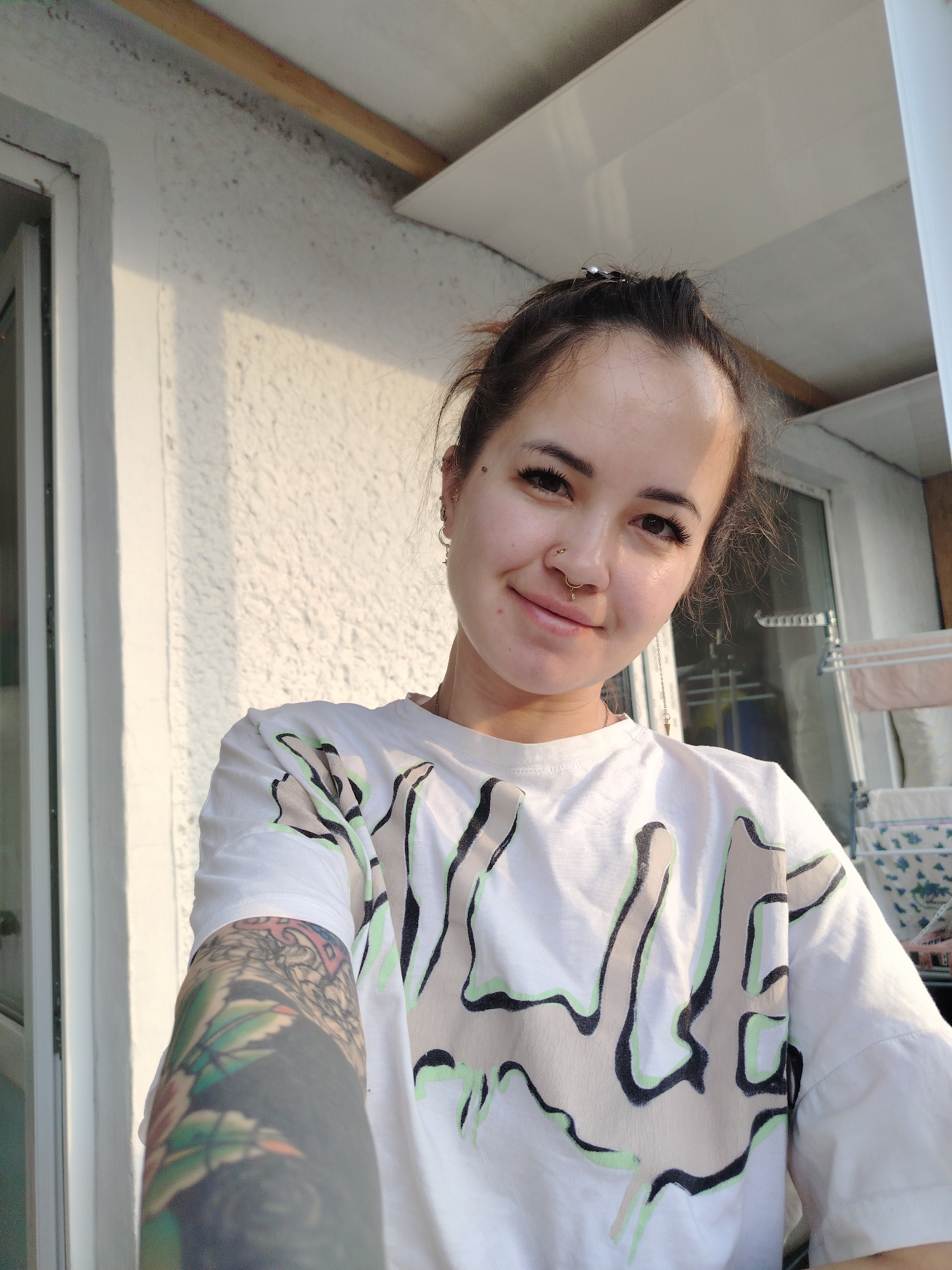 A young woman is taking a selfie outdoors. She has brown hair pulled back in a bun, pierced ears, and a nose ring. She is wearing a white T-shirt with a stylized design on the front and has a tattoo on her left arm. The background shows a white wall with a window, and a partial view of what appears to be a balcony or outdoor area. The location is possibly in Noviy, Russia, based on the prompt.