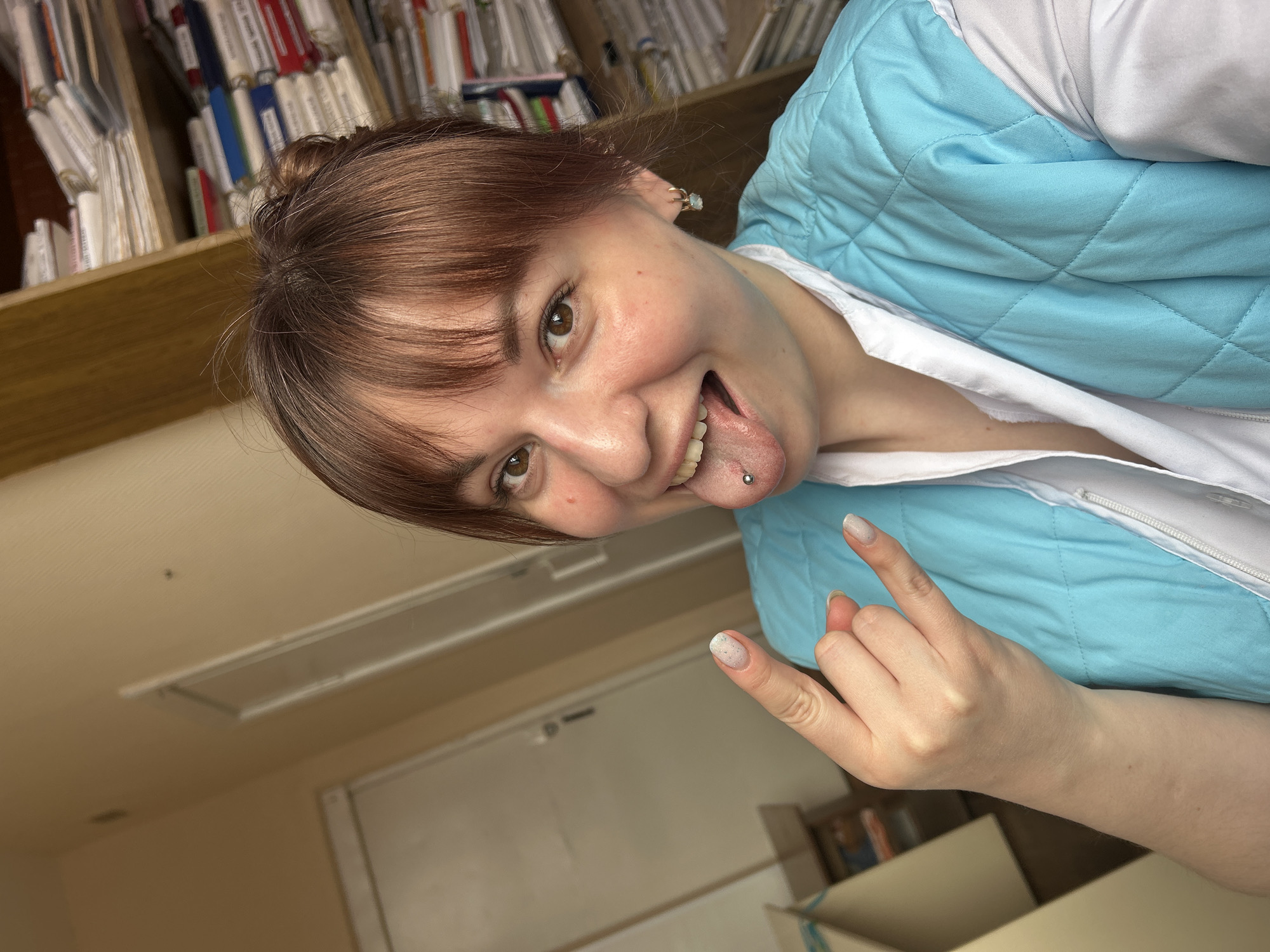 A young adult female is depicted in an indoor setting in the city of Foki, Russia. She has brown hair with bangs, brown eyes, and fair skin. She is wearing a white collared shirt under a light blue quilted vest. Her tongue is extended, revealing a silver barbell piercing. Her right hand is raised, forming a "rock on" gesture with the index and pinky fingers extended. Light-colored nail polish is visible on her fingernails. The background features light-colored walls, two white doors with trim on the left, and a wooden bookshelf filled with documents, folders, and binders on the right. A light-colored box is partially visible in the lower-left foreground. The individual is looking directly at the camera with a smiling expression.