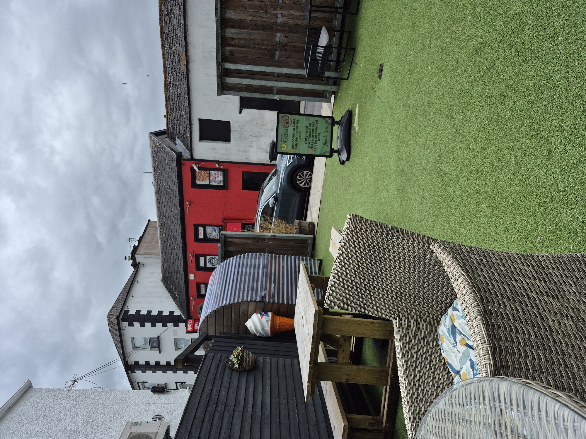 This image depicts an outdoor seating area, likely in Carrick-On-Shannon, Ireland, featuring artificial turf flooring. In the foreground, a wicker chair with a patterned cushion sits beside a glass-topped table. A wooden picnic table with benches is positioned further back. To the left, a dark wooden fence encloses the area, with a hanging basket containing greenery attached to it. An oversized ice cream cone decoration stands near a curved corrugated metal structure.
In the background, several buildings are visible. One building is painted bright red with black trim and features windows and signage, including "APACHE OPEN". Adjacent to it is a white building with black architectural details. A dark-colored car is parked on the street in front of these buildings. A green A-frame sign on the artificial turf reads "Kierans Gala Welcome to Gala Garden Seating area. Only food purchased on these premises can be consumed here. Thank You". Wooden fencing separates different sections of the outdoor space, and some potted plants are present. The sky is overcast with clouds.
