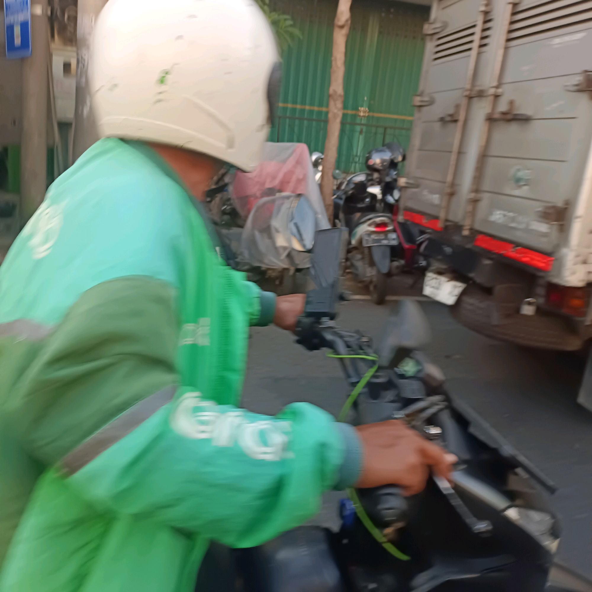 This is a motion-blurred, eye-level, outdoor shot of a person riding a motorcycle in what appears to be a city street, likely in Surabaya, Indonesia, judging by the context provided. The rider is wearing a white helmet and a bright teal and green jacket, possibly a uniform. The jacket has white lettering on the left sleeve. The rider's right hand is on the motorcycle's handlebar, operating the throttle. Behind the rider, to the right, is the side of a large, grey truck with horizontal slats and metal latches. To the left of the rider, there is a blue street sign with a white symbol. Several other motorcycles are visible in the background, some with red taillights. The environment suggests an urban setting with buildings and possibly a tree. The lighting indicates it is daytime.