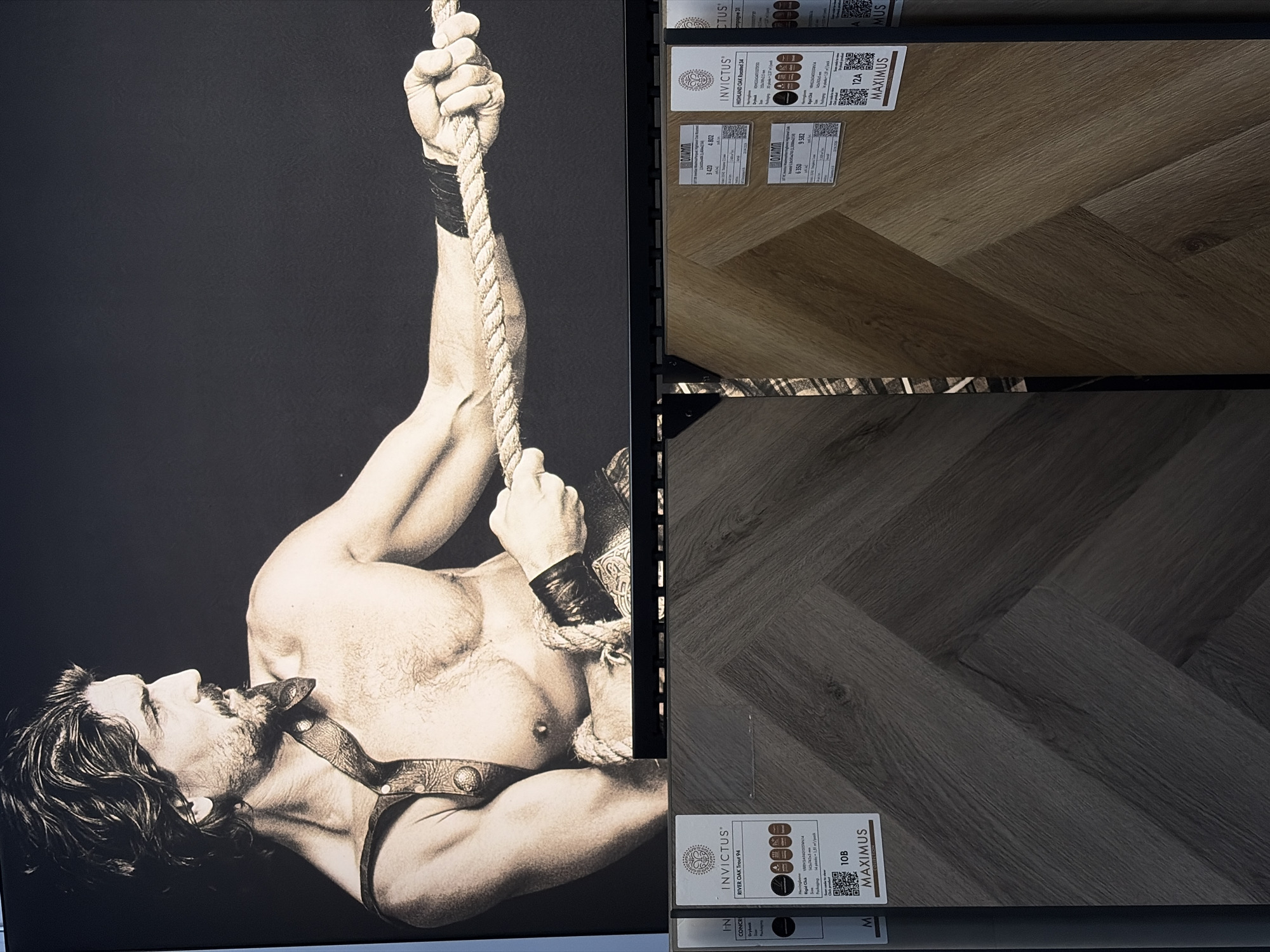 The image presents an advertisement for flooring materials, juxtaposed with an adjacent monochrome poster of a male figure. The man is depicted bare-chested, gripping a rope, and wearing a leather harness. The flooring samples display a herringbone pattern in light and dark wood tones, branded under the name "INVICTUS MAXIMUS" with additional details like "RIVER OAK Trout 94" and "HIGHLAND CAK Rocsted 34" included in the product descriptions. The scene appears to be a showroom or retail space. It's uncertain whether the location is Izhevsk, Russia, as the image lacks location-specific information.