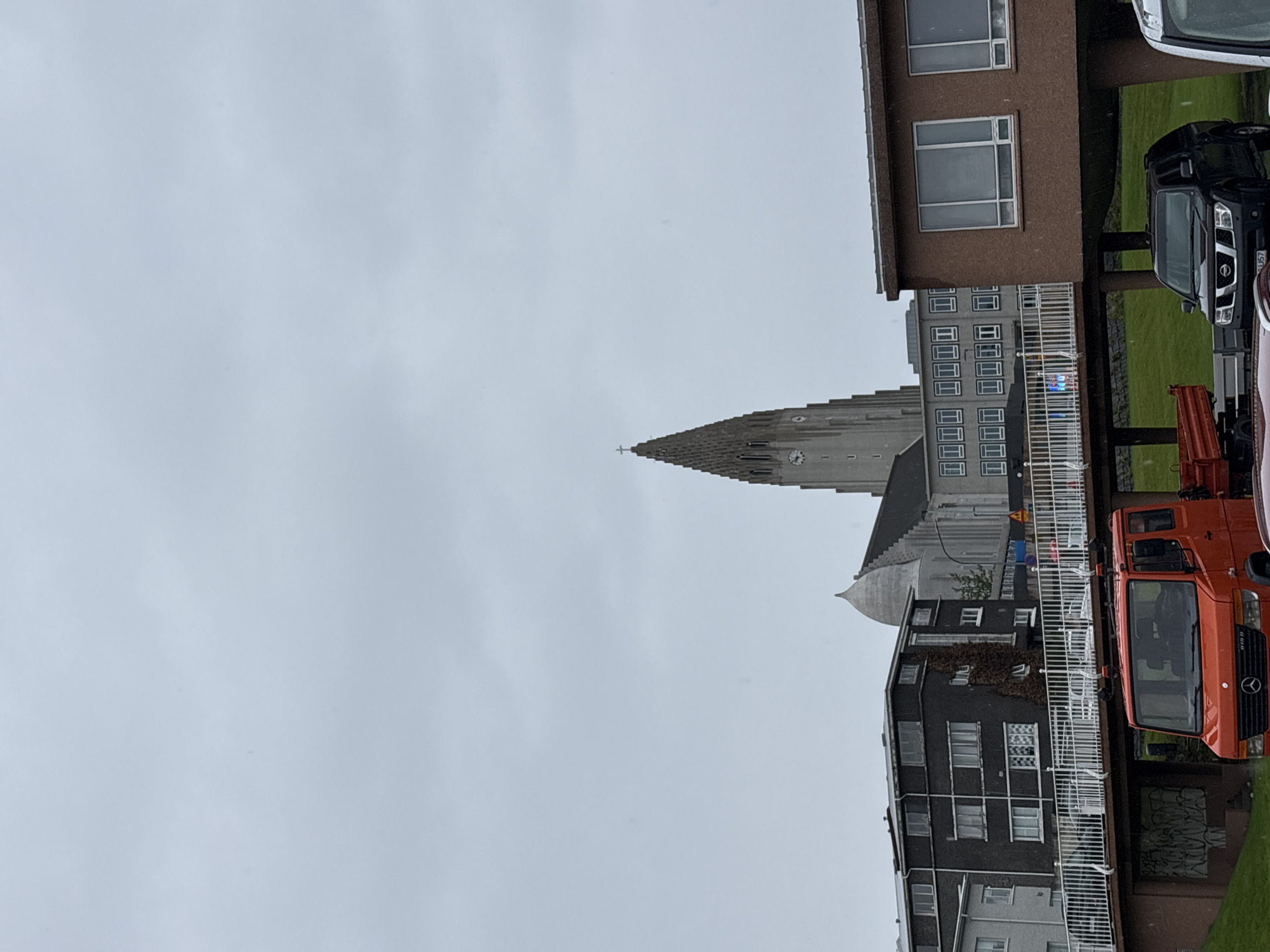 This is a frontal, eye-level, medium-shot photograph of Hallgrímskirkja church in Reykjavik, Iceland, taken on a cloudy day. The church, a prominent landmark with its distinct stepped, pyramidal tower and cross at the apex, dominates the background. In the mid-ground, several modern buildings with flat roofs and numerous windows are visible, including a dark gray apartment building and a lighter gray administrative building. A white railing runs along a pedestrian bridge, with a few cars visible driving on the bridge. In the foreground, an orange Mercedes-Benz truck is parked on a grassy area, and a black Nissan pickup truck is parked next to it. The sky is overcast and light gray, suggesting an overcast or rainy day, with a few visible raindrops on the lens and in the air. The overall scene depicts an urban environment with a mix of modern architecture and a significant historical and religious structure.