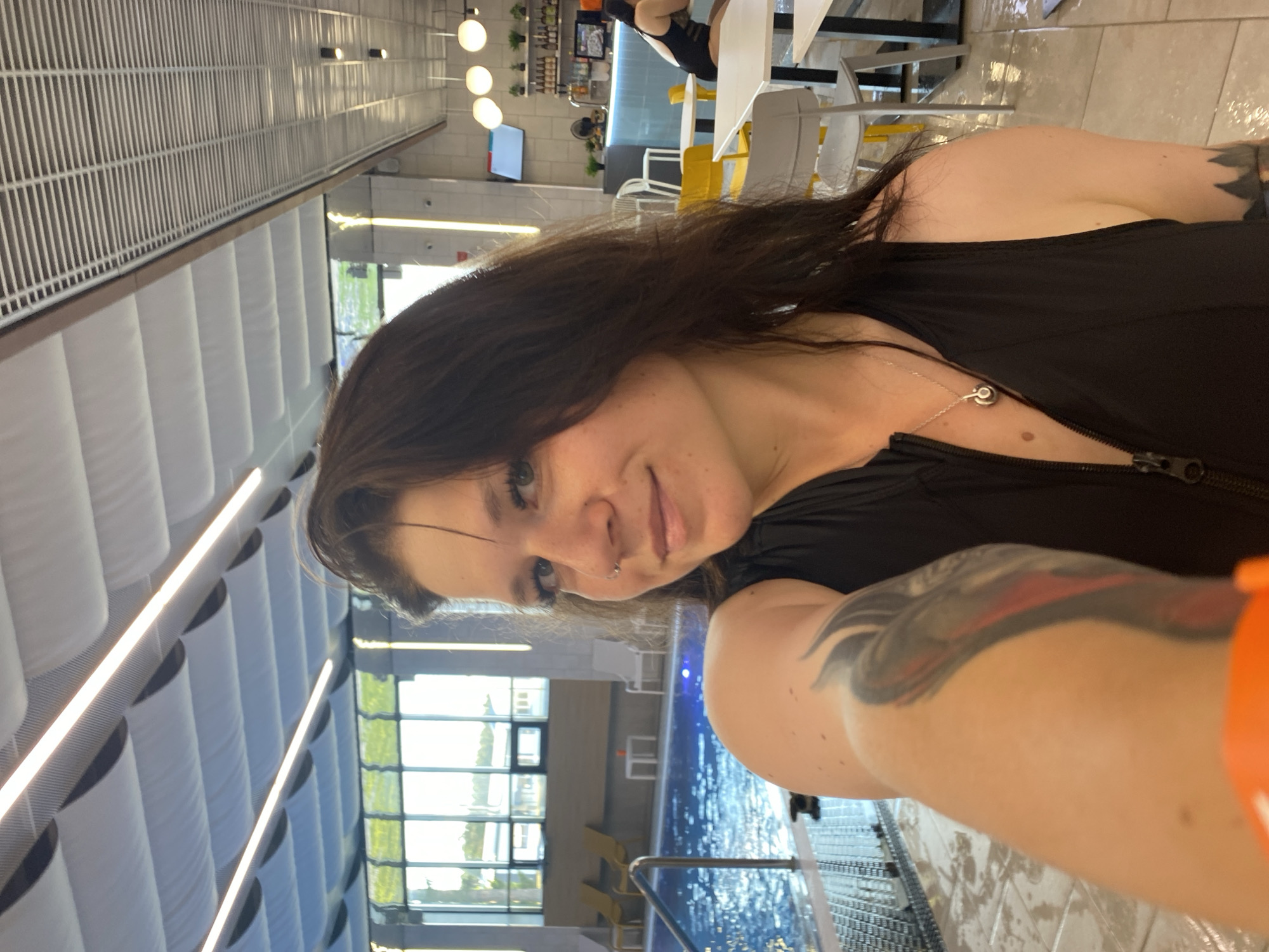 A young woman with dark hair and makeup is taking a selfie by a swimming pool. She is wearing a black swimsuit with a zipper. The pool is indoors with a glass ceiling and natural light filtering in. In the background, there are lounge chairs, tables, and a bar area. The location appears to be a spa or recreational facility.